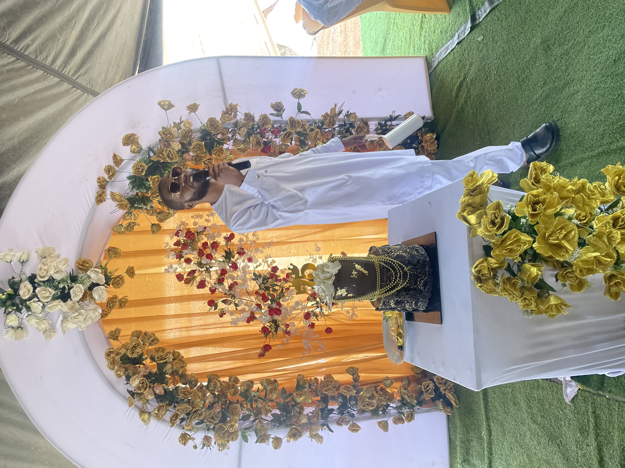A man wearing sunglasses and a white outfit is speaking into a microphone in front of a table with a decorated cake on it. The background features a decorated arch with flowers and fabric, suggesting an event or celebration. The location may be Jos, Nigeria, although this is not discernible from the image alone. The scene suggests a celebratory event with a speaker and a cake.