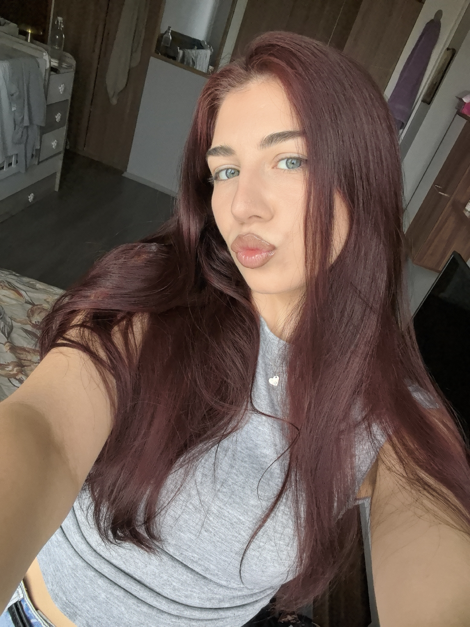 A close-up selfie shows a young woman with long, straight, reddish-brown hair and striking light blue eyes. She looks directly at the camera, her lips pursed in a playful pouting or kissing gesture, highlighted with a glossy finish. Her eyebrows are well-defined, and she has fair skin.
She is wearing a light grey, short-sleeved cropped top and a silver necklace with a small heart-shaped pendant. A faint, unidentifiable letter or symbol is visible on the pendant.
The setting is indoors, likely a bedroom or living area, in Stara Zagora, Bulgaria, captured during the daytime with natural light. The background reveals a light-colored dresser with multiple drawers and decorative handles on the left, topped with various items, including a small glass bottle. A patterned bedspread or throw in muted tones is visible in the lower-left. Dark wooden furniture or walls are seen further back, with a dark screen (possibly a television) partially visible on the right. The flooring appears to be dark wood or laminate. The woman's expression conveys a confident and engaging mood.