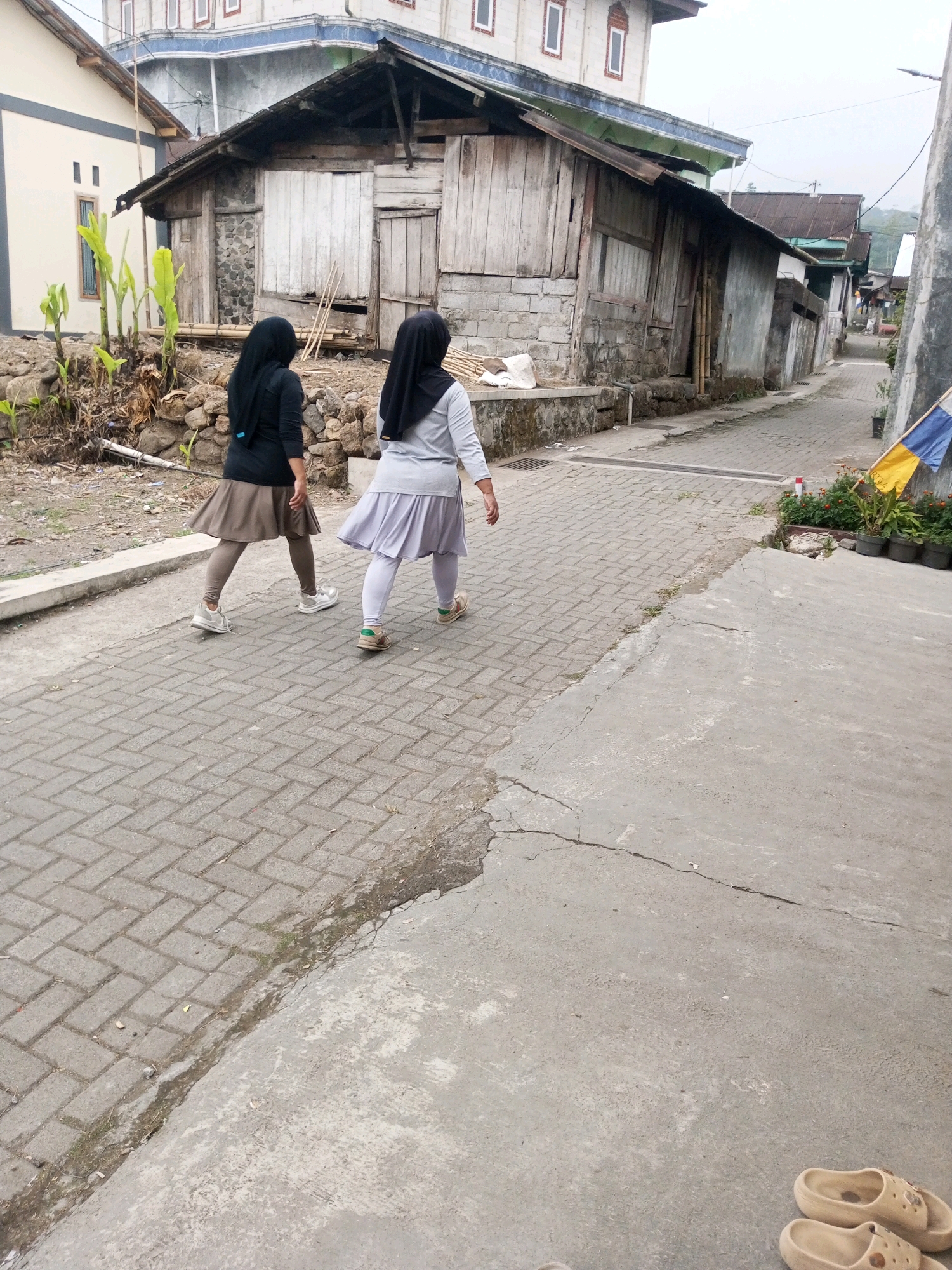 The image captures a scene in Andongsili, Indonesia, featuring two individuals walking away from the camera down a paved pathway lined with buildings. The pathway is constructed of interlocking grey bricks. To the left of the pathway, there is a dirt area with rocks and vegetation, adjacent to a weathered wooden building with a corrugated metal roof. Further down the pathway, the buildings appear more modern, with one structure displaying a blue and yellow flag. The ground is a mix of cracked concrete and brick paving. In the foreground, on the concrete area, a pair of light brown sandals are visible. The sky is overcast, and the overall impression is of a modest, possibly rural or semi-urban, neighborhood.