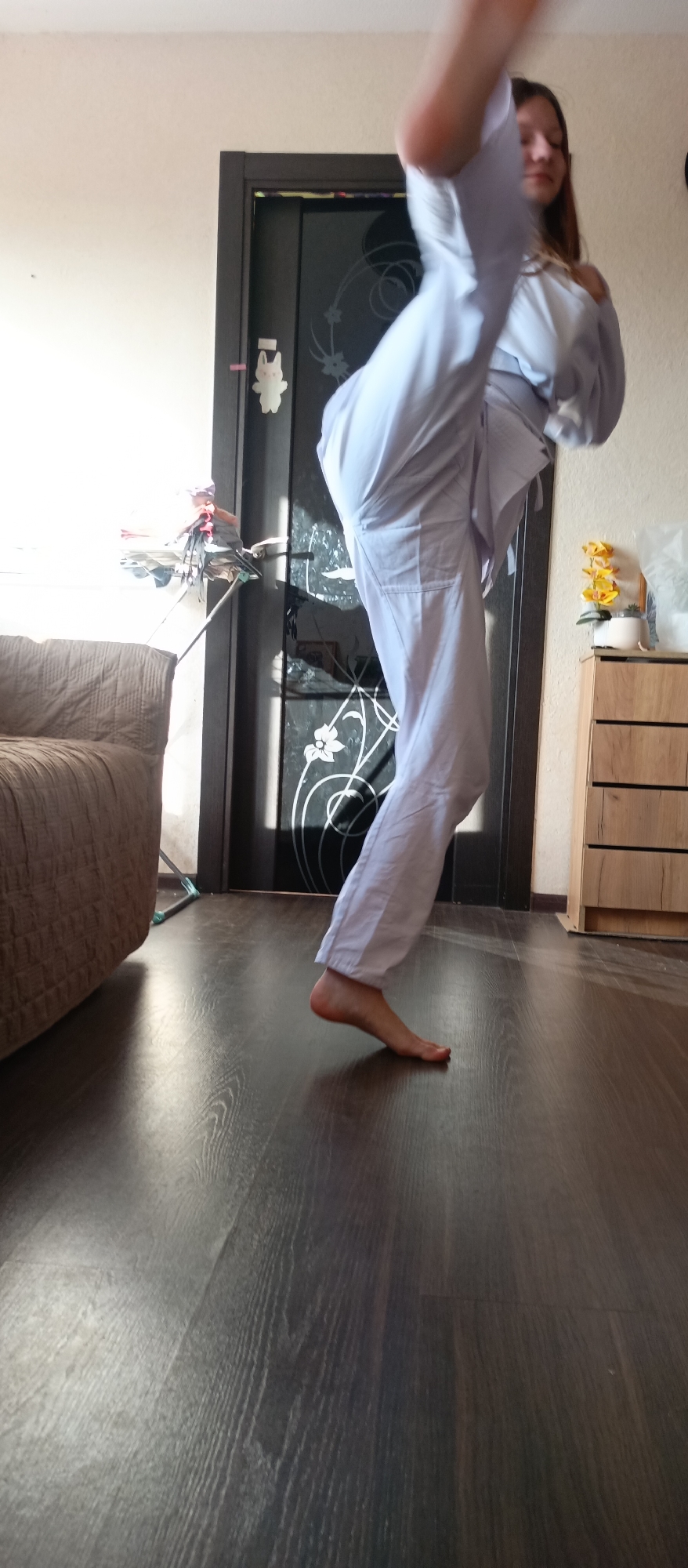 A young woman wearing a white martial arts uniform is practicing a high kick in a room. The room has dark wood flooring, a tan sofa, a brown door with decorative glass, and a small wooden cabinet. The setting appears to be inside a residence.