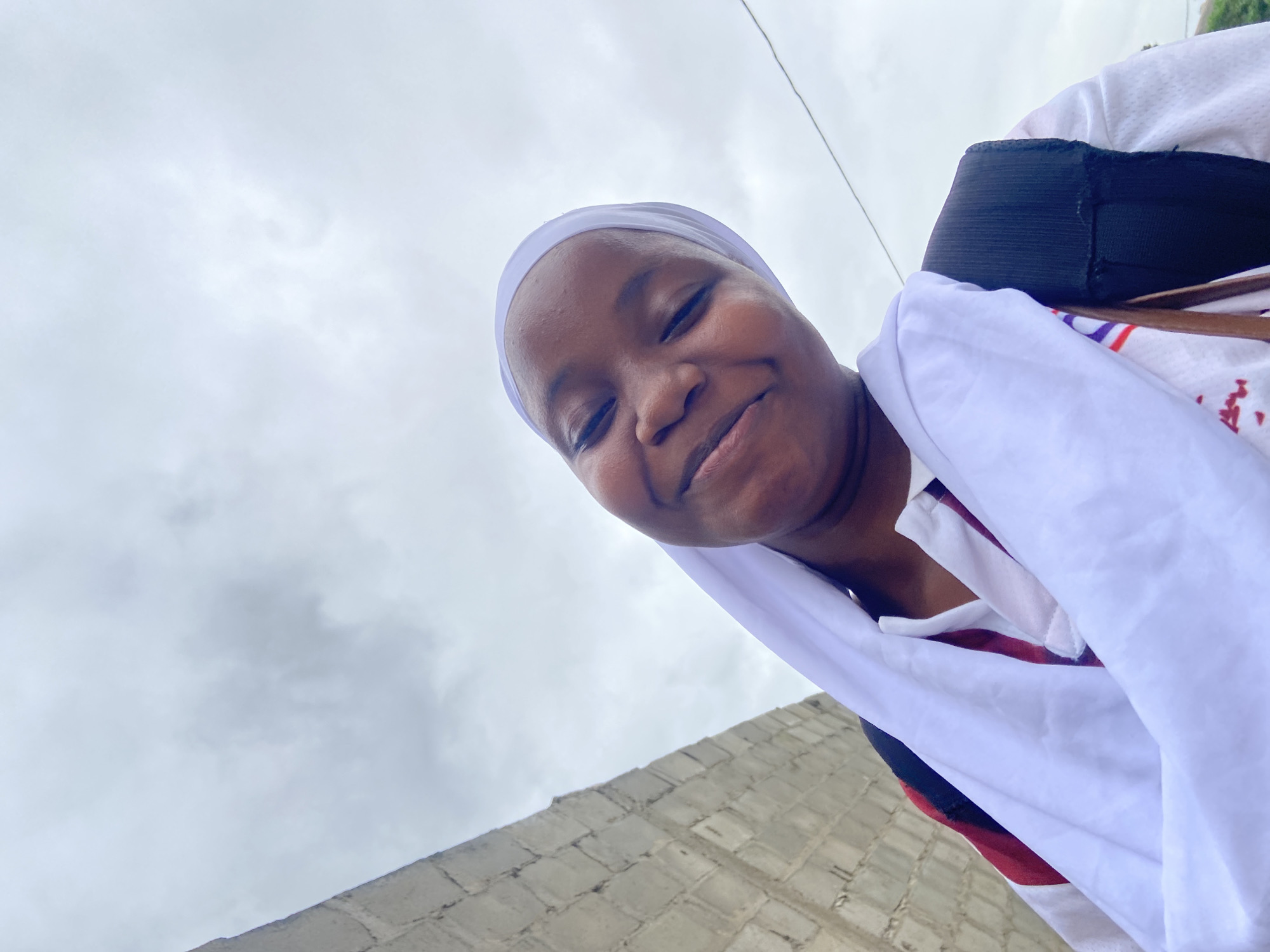 A young Black person wearing a white hijab and a white and red striped shirt is captured in a low-angle, close-up selfie. They are smiling and looking towards the camera. A dark backpack is visible over their shoulder. To the left of the frame, a wall constructed of light-colored concrete blocks extends upwards. The background is dominated by a cloudy, overcast sky. The scene is set outdoors, implied to be in Egan, Nigeria, given the context.