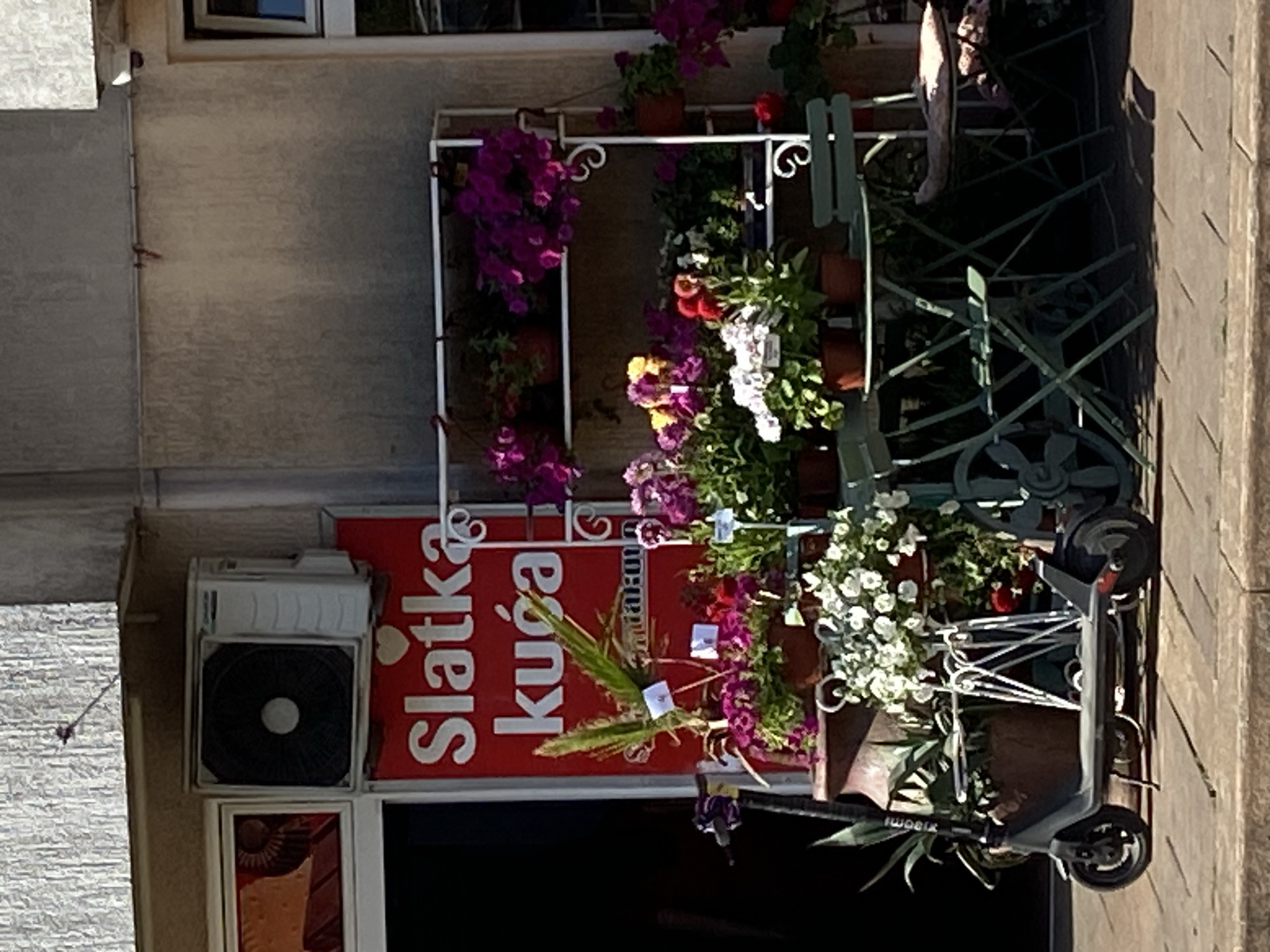 A street-level view reveals a portion of a building with a shop facade. The shop sign reads "Slatka kuća" in white lettering against a red background. An air conditioning unit is mounted above the shop front. Several potted plants in a variety of colors are displayed in front of the shop, supported by a white metal rack. Green metal chairs and a gray Xiaomi scooter are positioned on the sidewalk in front of the plants. The setting is outdoors, on a paved street. This is Budva, Montenegro.