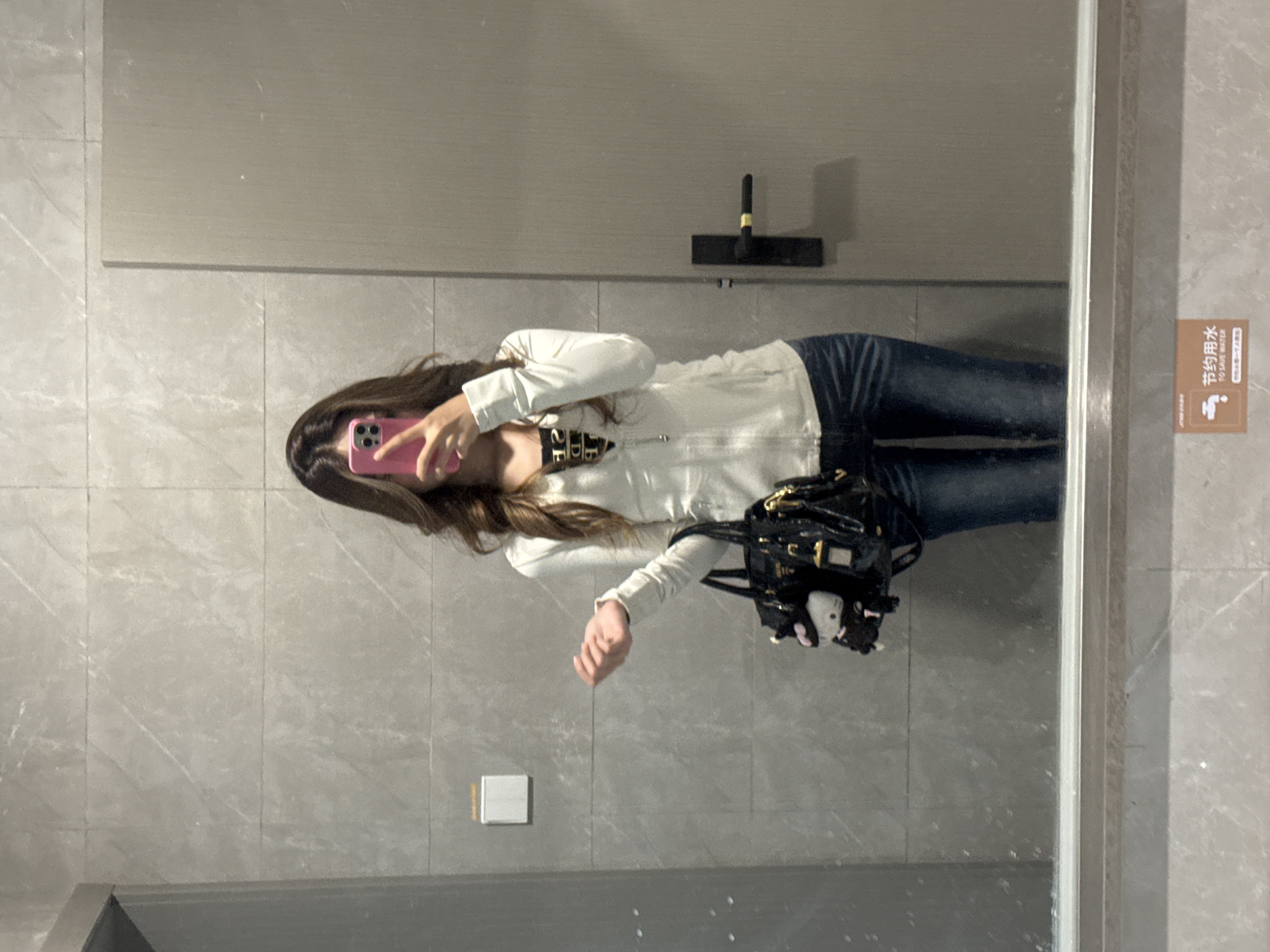 A woman is taking a mirror selfie in a public restroom. She is wearing a white collared shirt and jeans, and has a black purse over her shoulder. The restroom has gray tile walls and a mirror with a sign about water conservation in Chinese. The location is Nan'an District, China.