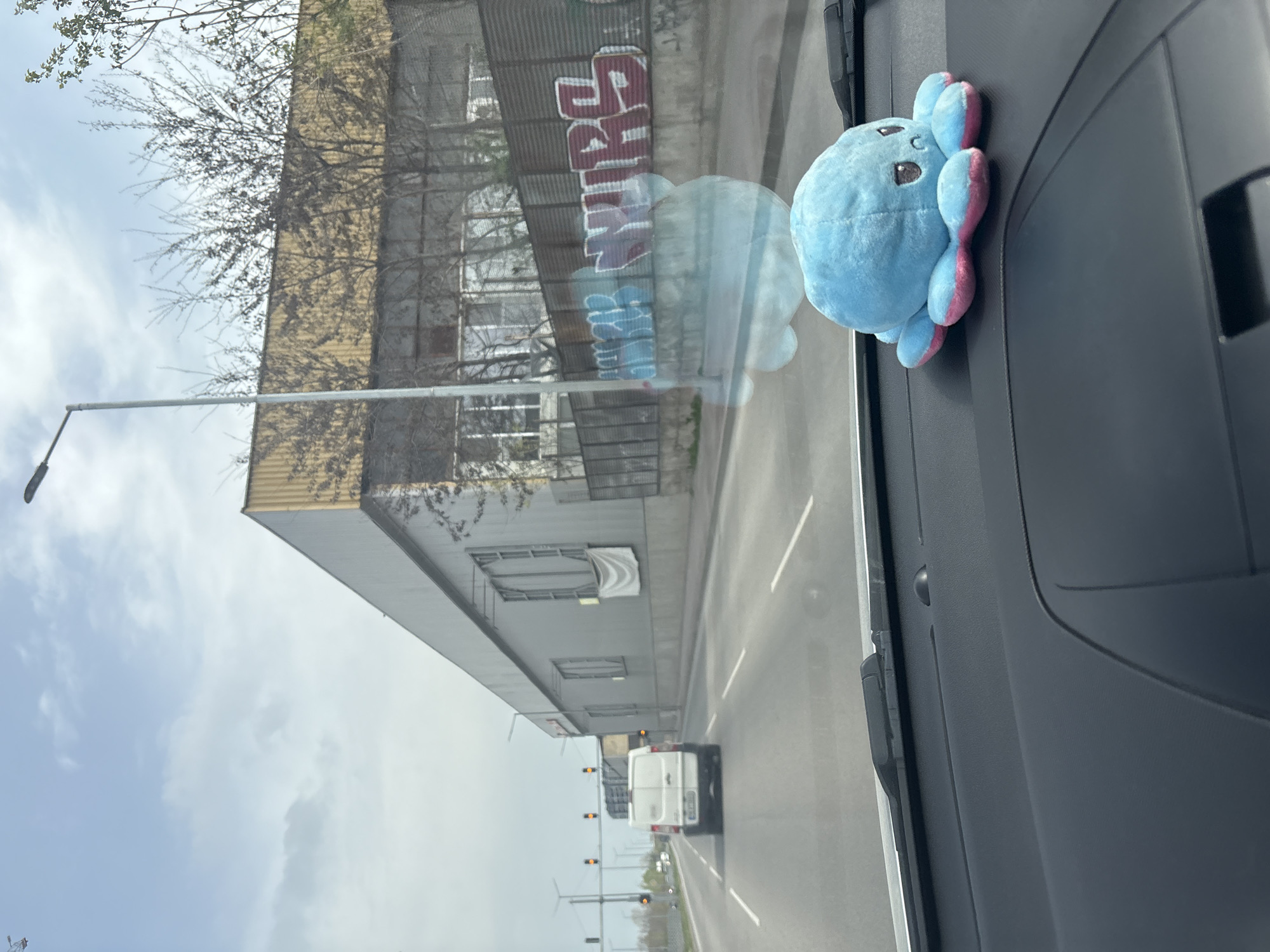 The photograph appears to be taken from inside a vehicle, likely a car or truck, looking through the windshield towards the exterior environment. A light-blue octopus plush toy with pink accents is attached to the right side of the dashboard or windshield. The external view shows a city street with an industrial building to the left. The building is multi-storied and shows signs of wear and aging, with visible graffiti on the upper part. A street light post is also visible in front of the building. In the distance, a white van travels on the road. Traffic lights are visible in the distance above the van. The sky is overcast, and the city location is Unknown, Unknown.