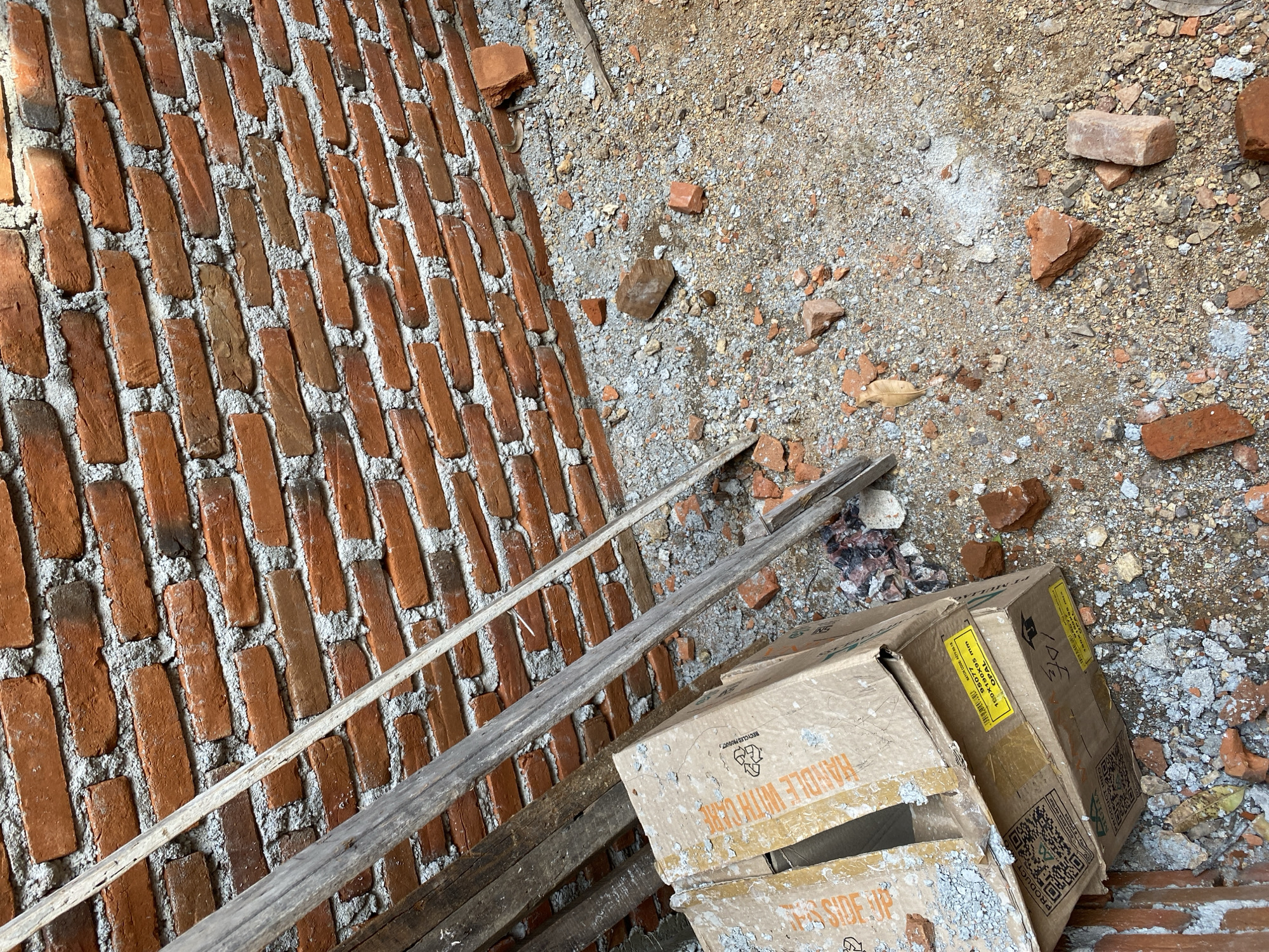 This is a high-angle, eye-level medium shot of a construction site. The scene is dominated by a red brick wall on the right and in the background, with cement mortar visible between the bricks. In the foreground, there is loose gravel and dirt, with scattered red brick fragments. To the left of the frame, two cardboard boxes are stacked, one on top of the other. The top box is open, and both have tape securing them. Several wooden planks are leaning against the wall and the boxes. The lighting suggests it is daytime. The location is unknown.