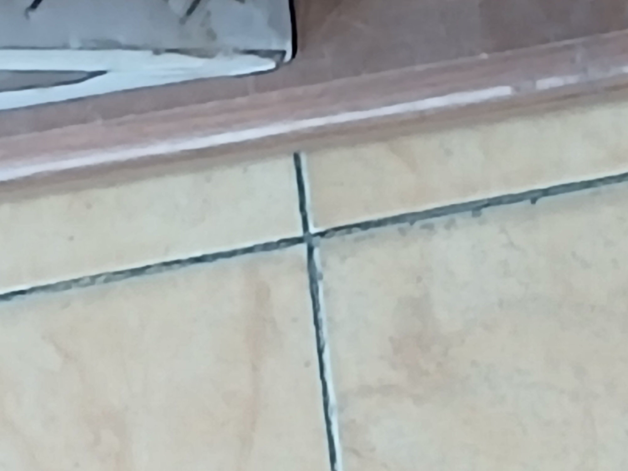The image shows a close-up view of a tiled floor with grout lines. To the right, a wooden frame or doorjamb is visible, with a white object partially obscured behind it. The scene appears to be indoors, possibly a residential or commercial space in Juan Griego, Venezuela. There are no people or animals in the frame, and no apparent actions or interactions are taking place. The lighting is moderate, and the overall condition of the floor and surrounding objects appears to be ordinary.