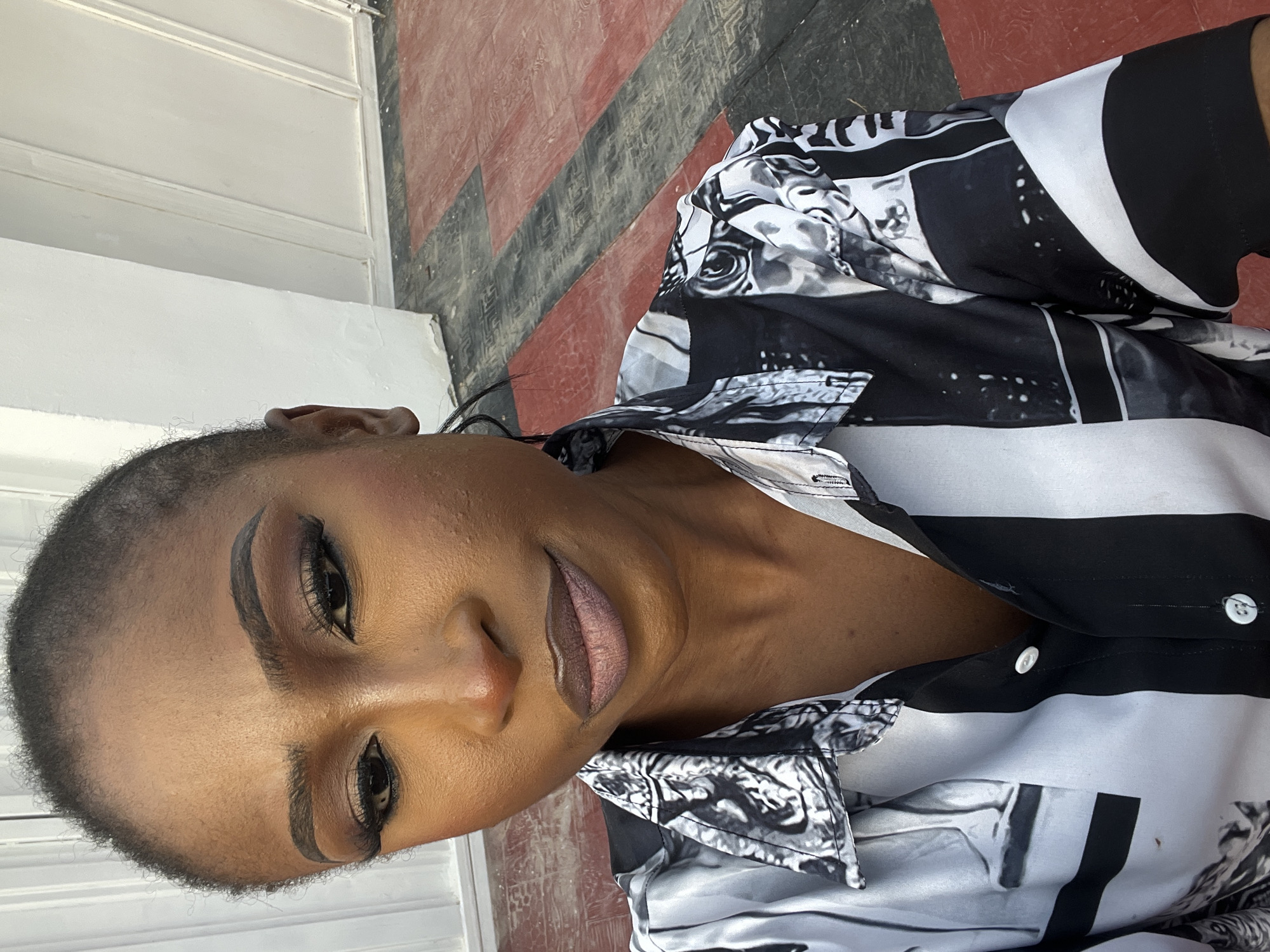 The image is a close-up, high-angle selfie of a Black woman posing outdoors. She has her hair pulled back and is wearing makeup, including eyeshadow and lipstick. She is wearing a black and white patterned button-down shirt. The background shows a textured floor with red and black tiles and a white wall. The scene appears to be in Abuja, Nigeria, based on the provided context.