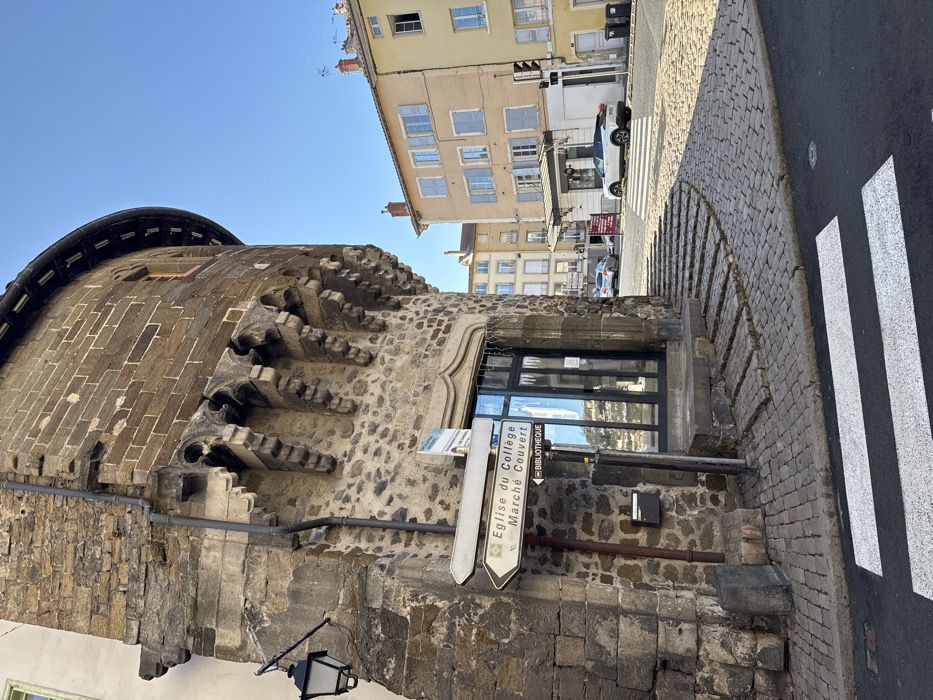 The image depicts a street scene in Le Puy-en-Velay, France, characterized by historical architecture and urban infrastructure under a clear blue sky.
In the foreground, a white pedestrian crossing with two thick stripes spans an asphalt road. Adjacent to this, on the left, is a prominent, multi-story stone building with a rounded, fortified-style corner section. The lower facade consists of rough-hewn stone blocks, transitioning to more regular ashlar masonry on the upper levels. A black, decorative street lantern is affixed to its left-hand wall, and a grey downspout runs vertically along the building's corner. Below a decorative stone architrave, a modern entrance features a dark-framed glass door. To the left of this door, a brown metal pole holds two white directional signs. The top sign, pointing left, reads "Eglise du Collège." The bottom sign, pointing right, indicates "Marché Couvert" and "BIBLIOTHEQUE." A black wall-mounted mailbox is positioned below these signs. Curved stone steps lead from the cobblestone sidewalk up to the building's entrance. Above the doorway, the rounded building section is supported by a series of intricately carved stone corbels, topped by a section of lighter stone blocks and a small, rectangular window with a red-brown frame.
The cobblestone street curves to the right, leading towards an intersection. A white SUV is parked beyond the pedestrian crossing on the right side of the street, facing away from the viewer. A black traffic light pole stands on the right sidewalk. In the background, several traditional multi-story European buildings are visible, including a cream-colored structure with light blue shutters on its windows and a light yellow building with similar architectural features and red-tiled rooftops. No individuals are present in the scene. The ambient light suggests daytime conditions.