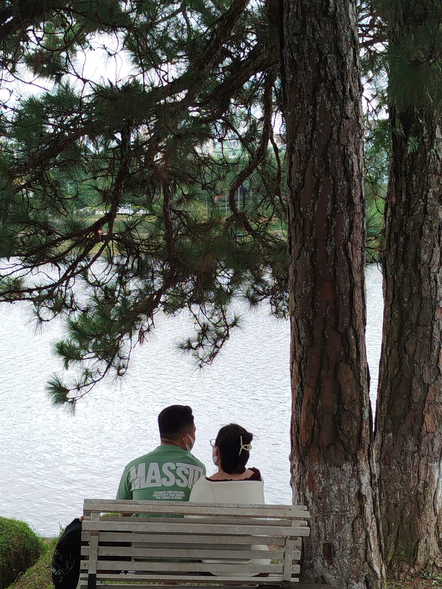 Two individuals, a man and a woman, are seated on a wooden bench facing a body of water. The man, on the left, wears a green long-sleeved shirt with white lettering and a white face mask. The woman, on the right, has dark hair styled with a clip and wears a white top, also with a white face mask. They are positioned under the canopy of pine trees. The background shows the calm surface of the water, with faint hints of buildings and greenery along the distant shore. The scene appears to be a park-like setting by a lake, commonly found around Xuan Huong Lake in Da Lat, Vietnam, offering a peaceful vista. The foreground is dominated by the textured bark of two large trees and the wooden bench.