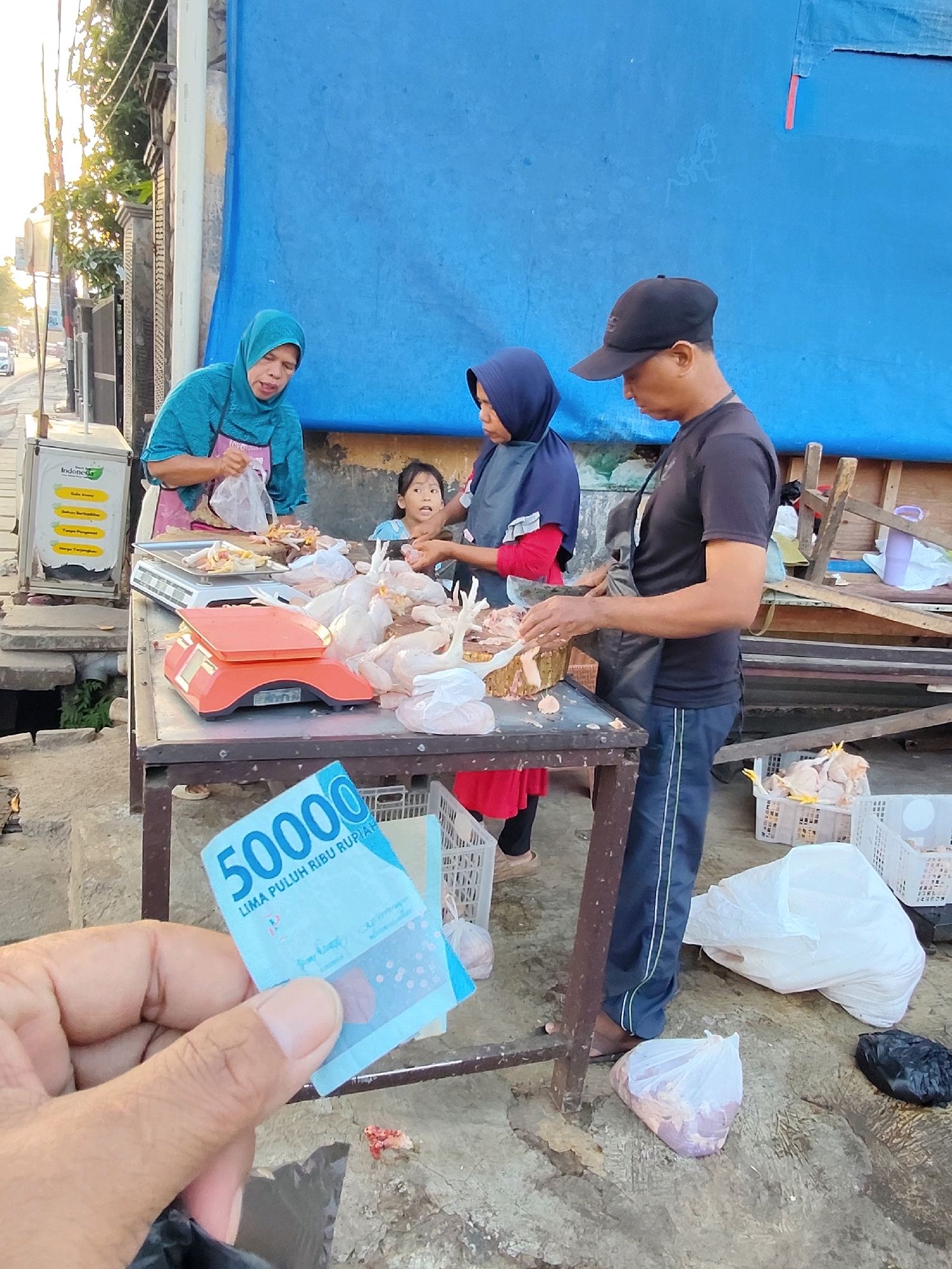 The image depicts a street market scene in Purwakarta, Indonesia. In the foreground, a hand holds a 50,000 Indonesian Rupiah banknote. Behind it, a table is set up with a scale and raw chicken for sale. Three people are visible. A woman wearing a blue hijab and apron is attending to the chicken. A young girl in a blue hijab stands next to her, looking at the camera. A man wearing a black cap and t-shirt, likely a vendor, is preparing the chicken. In the background, a large blue tarp serves as a backdrop, and various crates and bags of goods are scattered around the market stall. The environment appears to be outdoors, with sunlight suggesting it is daytime.