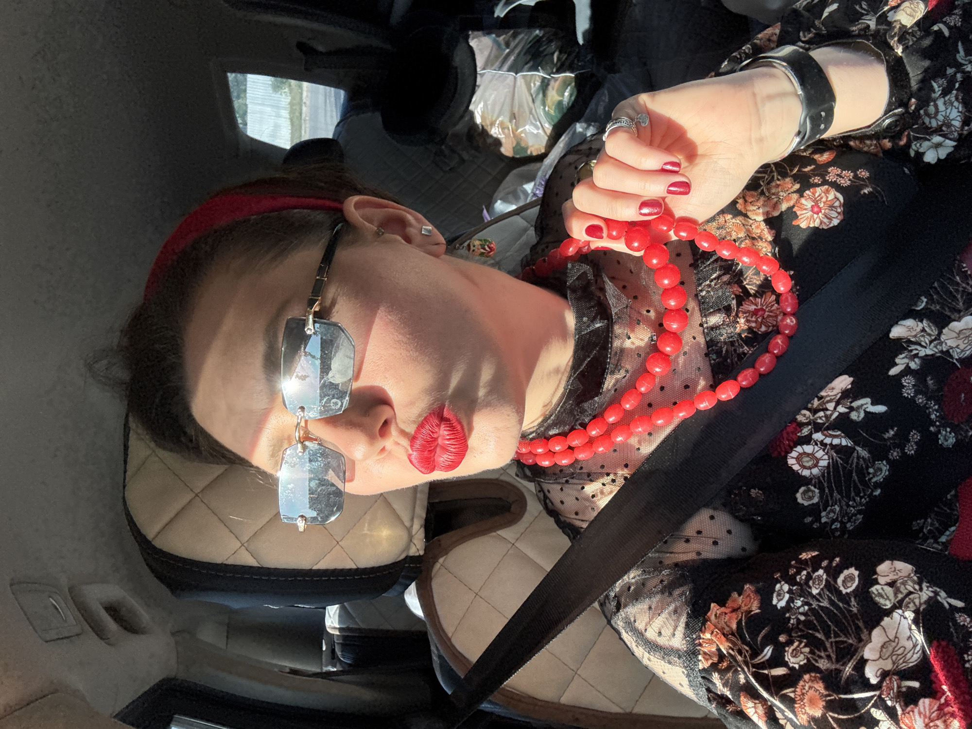 A woman is seated in the passenger seat of a car. She is wearing a red headband and glasses. She has red lipstick on. She is wearing a black top with floral print and a mesh underlay. She is holding a red beaded necklace. She is wearing a black cuff bracelet and a ring. She is wearing a seatbelt. The headrest of the car seat behind her is tan with a quilted pattern. The location is not discernable based on the given image.