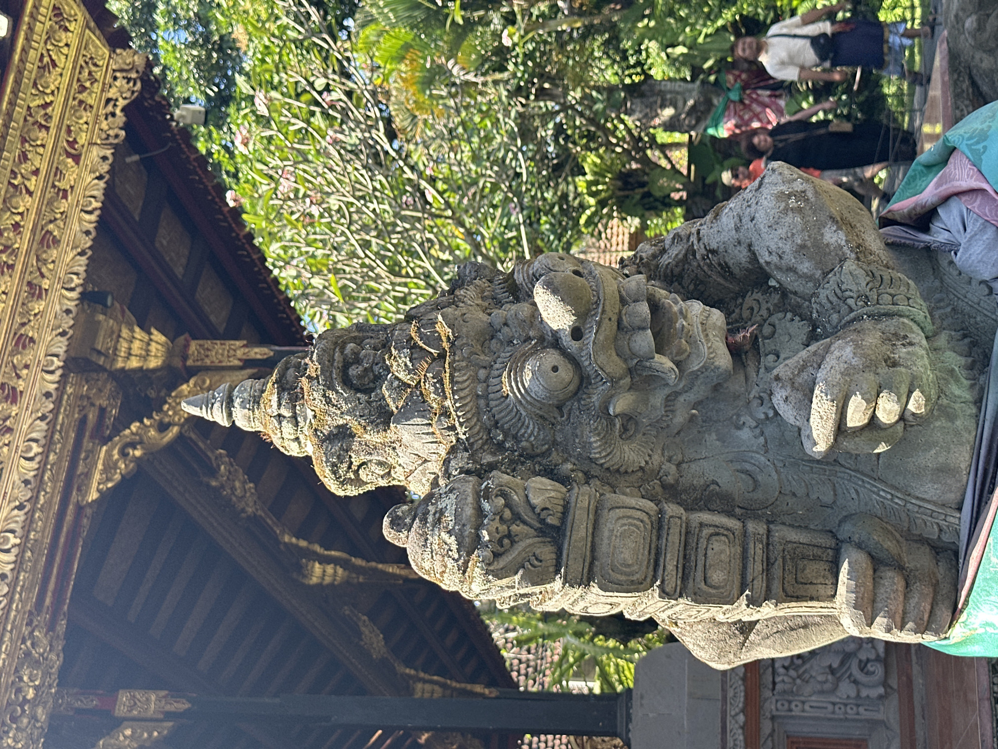 In Ubud District, Indonesia, a weathered stone statue of a mythical creature stands prominently in the foreground. The statue, covered in moss, features a fierce expression with wide eyes, an open mouth revealing teeth, and a towering headdress. It is adorned with a green sash around its lower body. Behind the statue, a traditional Balinese temple structure with intricate golden carvings and a wooden roof is visible. Lush green foliage, including trees and flowering plants, surrounds the temple. In the background, several people are walking along a path, some wearing traditional Balinese attire, indicating this is likely a tourist or cultural site. The lighting suggests it is a sunny day.