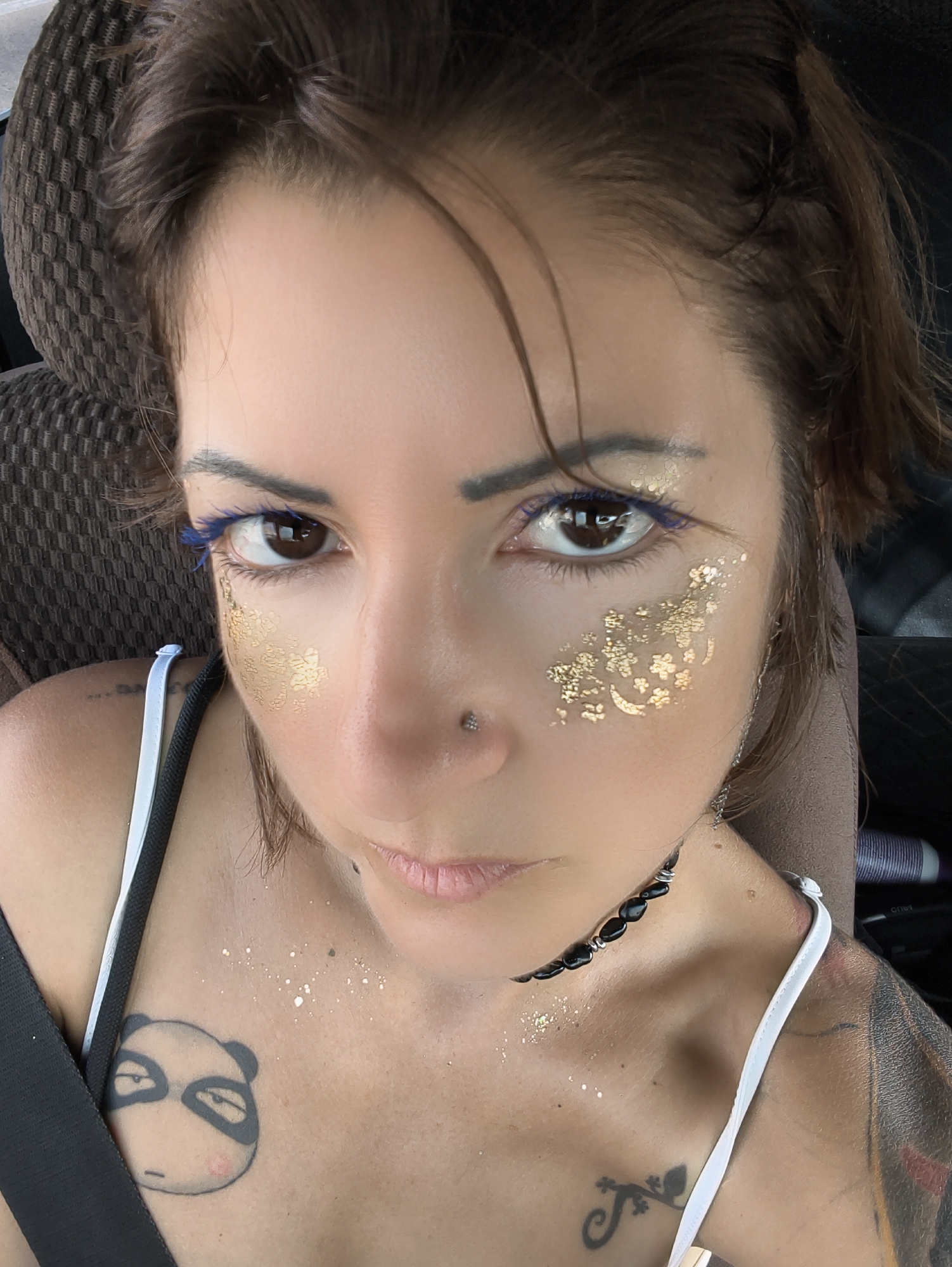 A close-up, selfie-style portrait of a woman is captured, focused on her face and upper chest. The woman has brown hair, styled in loose waves. She is wearing makeup, including blue mascara and gold glitter accents on her cheeks and forehead. She has a nose ring. Visible on her chest are two tattoos: a panda face on the left and a stylized lizard on the right. She is wearing a black necklace with black beads and what appears to be a black seatbelt crossing her chest. The background is blurred, suggesting an indoor or vehicular setting. The lighting is bright, creating some highlights on her skin and the glitter. The image quality is good, with clear detail on the subject's face and tattoos. The location in Ramat Negev Regional Council, Israel, is not discernible from the visual content of the image itself.