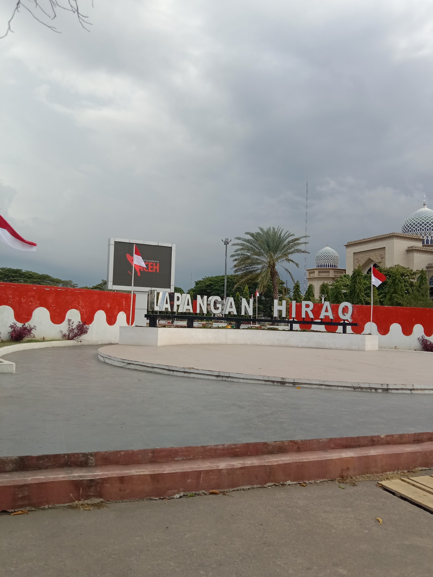 The image depicts the "LAPANGAN HIRAQ" (Hiraq Field) in Lhokseumawe, Indonesia. In the foreground, there is a paved area with a low, red-and-white decorative wall. Large, three-dimensional white letters spelling out "LAPANGAN HIRAQ" are prominently displayed on a white platform. Two Indonesian flags are visible, one a large white and red flag waving in the upper left corner, and another smaller flag planted near the "HIRAQ" sign.
Behind the sign, a large digital billboard displays a red star and text in a language that appears to be Indonesian, with "ACEH" visible at the bottom. A few small green plants are situated in front of the red-and-white wall. To the right of the sign, a palm tree stands tall.
In the background, a distinctive mosque with a blue dome and intricate architectural details dominates the skyline. A tall, slender tower or antenna is also visible in the distance. The sky is overcast with grey clouds, suggesting a possibly humid or rainy day. The overall scene captures a public space in Lhokseumawe, characterized by its national symbols, public signage, and notable religious architecture.