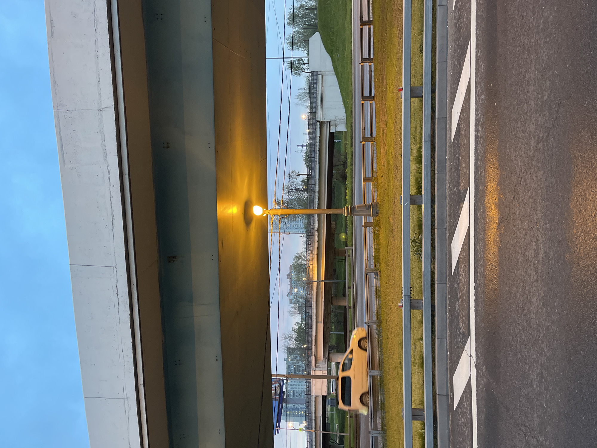 The image shows a view from underneath a bridge in Khimki, Russia. To the right, a highway runs along a green grassy area. A street light shines brightly between the highway and the bridge. In the background, through the gap between the bridge and highway, are other bridges and buildings. A car is moving along the highway. The sky is visible in the upper portions of the image and appears to be dusk or dawn.