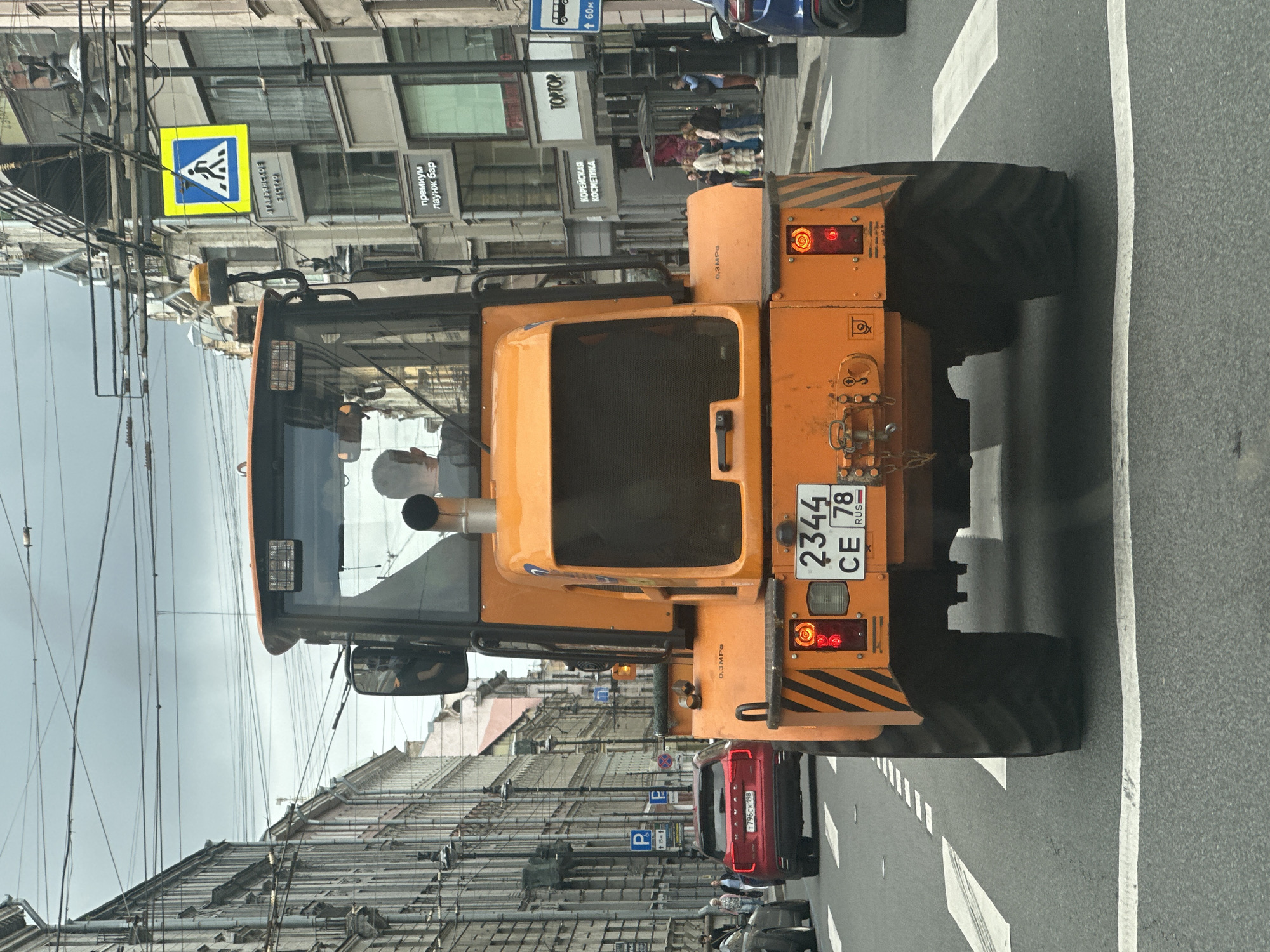 This is a rear view, medium shot of an orange construction vehicle, likely a backhoe loader, driving on a city street in Saint Petersburg, Russia. The vehicle has large rear tires and the back of its cab is visible, with a person's head seen through the window. A license plate with the numbers "2344 CE 78" and "RUS" is attached to the rear of the vehicle. The street is paved and marked with white lines, indicating it is a road with a pedestrian crossing. Buildings line both sides of the street, with ornate architecture typical of a historic European city. Overhead power lines crisscross the sky. Several other vehicles, including a red SUV and a dark-colored car, are also visible on the street. A pedestrian crossing sign is visible on a pole to the right of the vehicle. The sky is overcast.