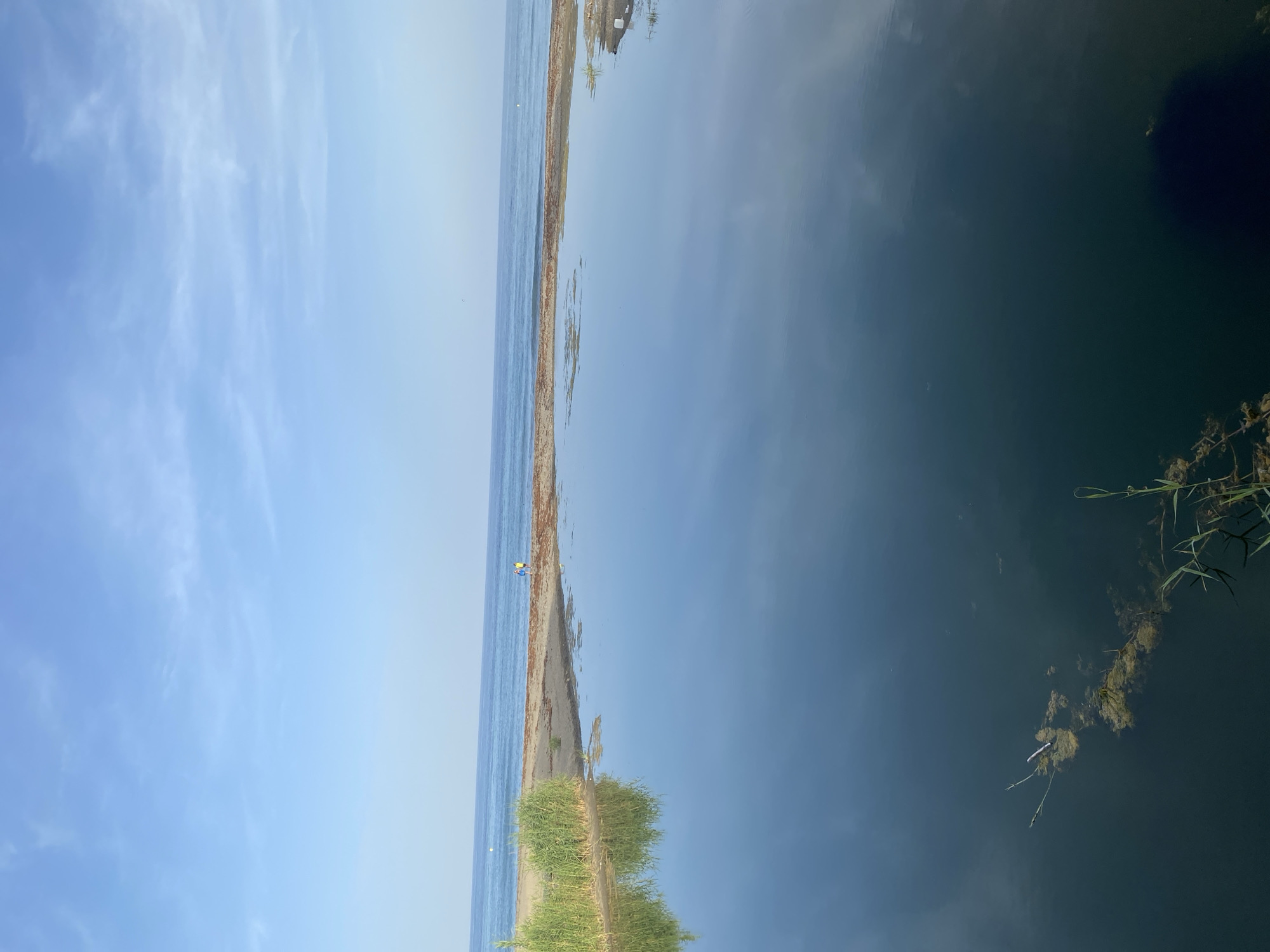 The image depicts a tranquil scene at the Resinera-Voladilla, Spain. A narrow strip of sandy shore separates a calm, deep blue body of water from a clear sky dotted with wispy white clouds. The water reflects the sky, creating a mirror-like effect.
On the shore, a small figure in a yellow top is visible, appearing to be a person. Further along the shore, green vegetation grows, its reflection also visible in the water. In the deeper water, some floating debris and aquatic plants can be seen. The lighting suggests it is daytime, possibly late afternoon, given the soft light and the clear, bright sky. The overall mood is peaceful and serene, with a sense of stillness and natural beauty. There is no visible text in the image.
