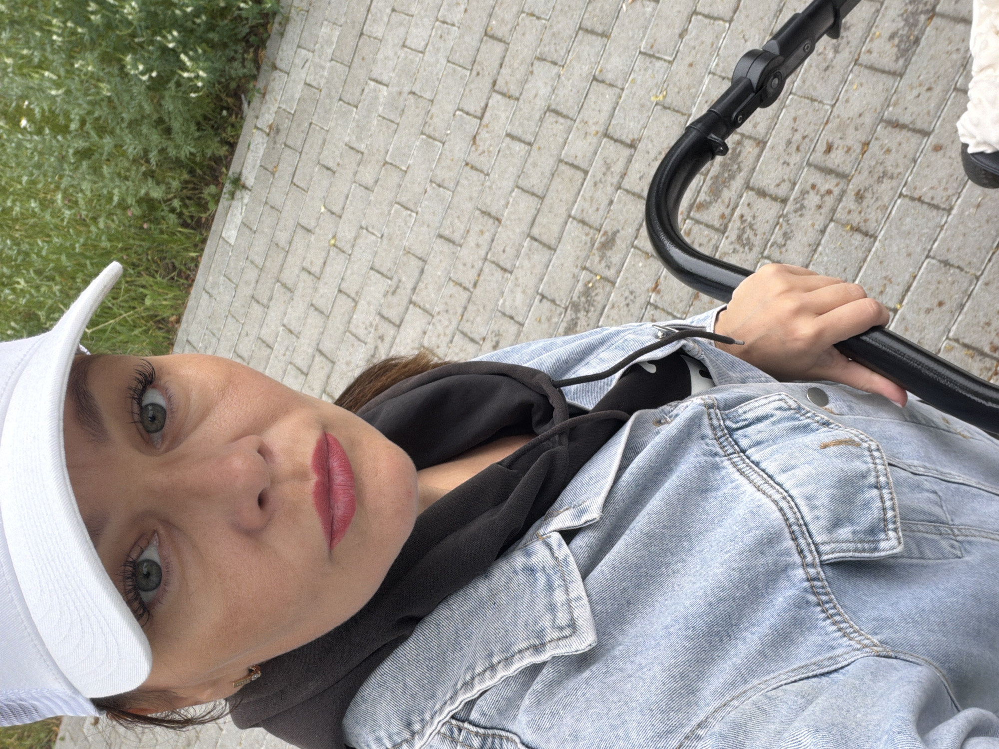 The media file features a selfie of a woman in Chaikovsky, Russia. She is wearing a light blue denim jacket, a black hoodie, and a white baseball cap. Her makeup includes noticeable mascara and pink lipstick. In the background, a paved walkway made of rectangular stones is visible, bordered by green vegetation. A black stroller handle is held by her right hand, suggesting she is either pushing or holding onto a stroller. The image appears to be taken during the daytime, with natural lighting. The overall mood seems casual and perhaps active, given the presence of the stroller. There is no visible text in the image.