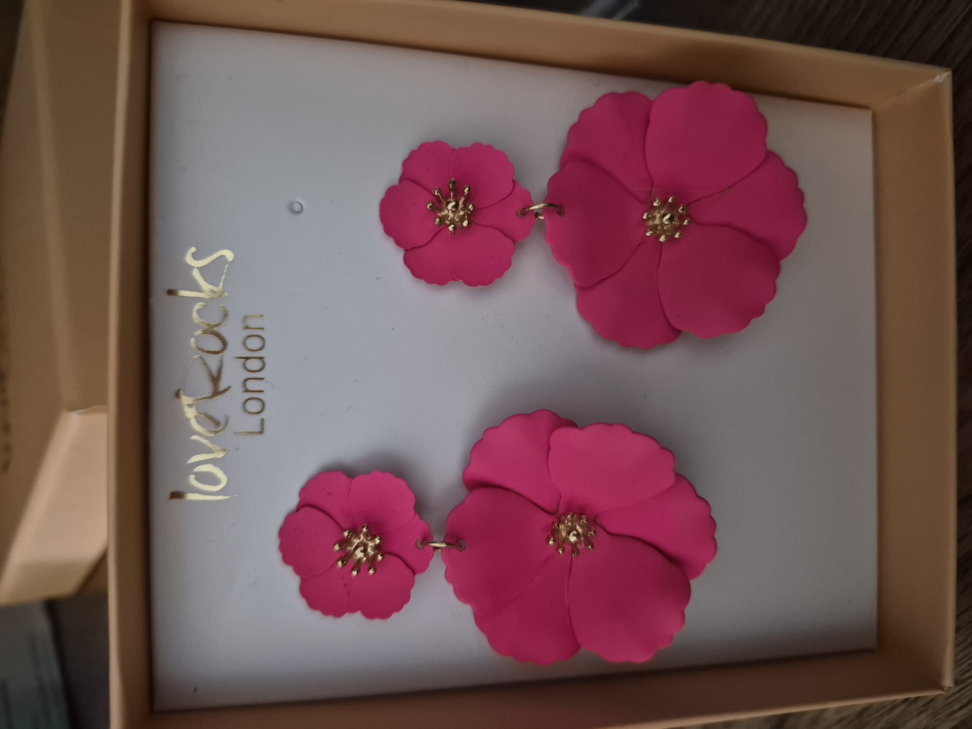 A pair of bright pink, double-layered flower-shaped earrings are displayed in a light grey box with gold lettering that reads "love rocks London". The earrings are hanging from their clasps, with the larger flower at the bottom and a smaller flower above it. Each flower has a gold center. The box is partially open, revealing the earrings inside. The background is blurred, but appears to be a wooden surface. The scene is well-lit and the colors are vibrant.
