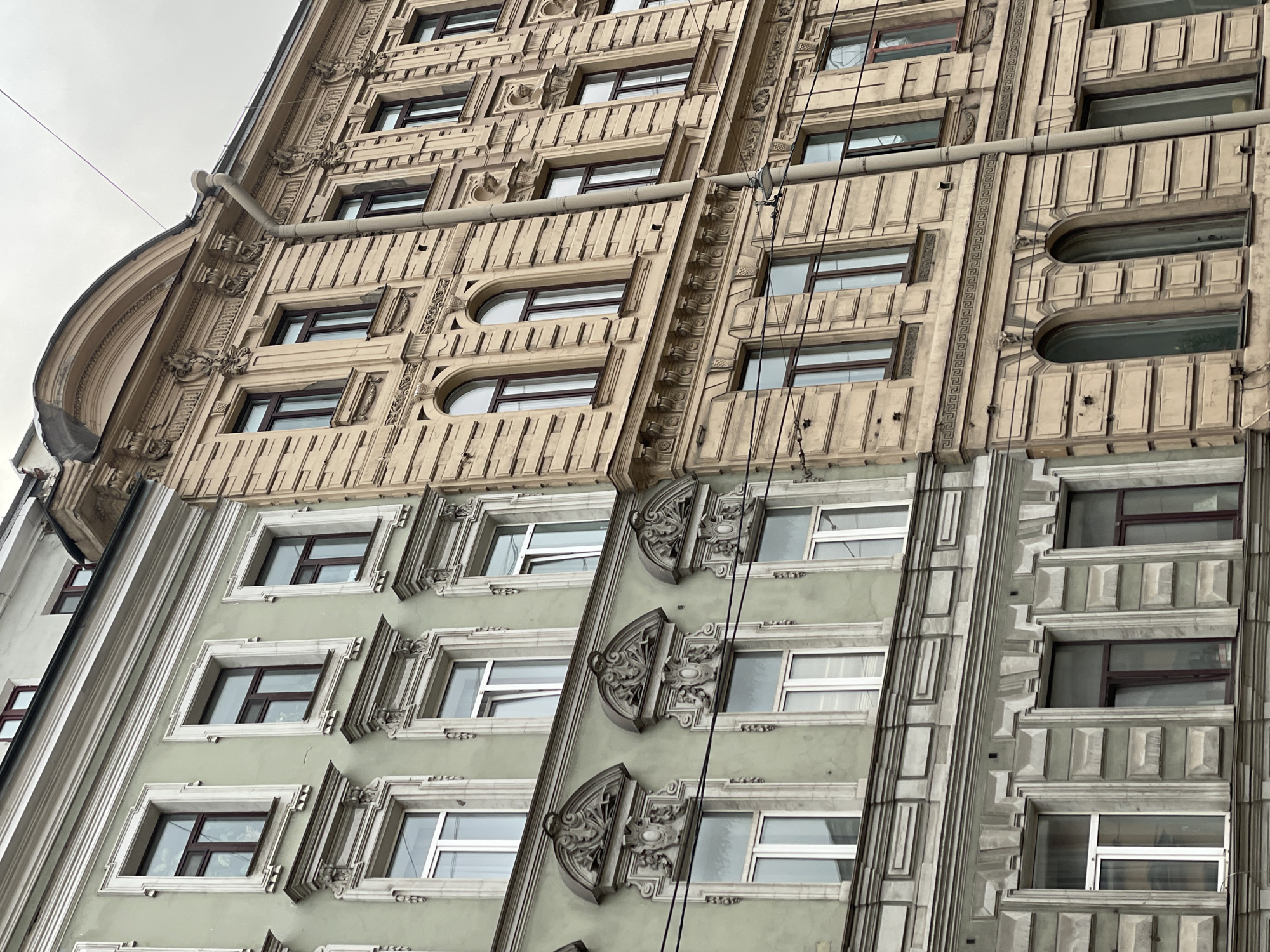 The image depicts the facade of a building in Saint Petersburg, Russia. The building's architecture is ornate, with decorative elements around the windows and along the vertical lines of the building. The building consists of at least three distinct sections, each with a unique color and design. The sections transition from a beige color on the upper floors to a light green in the middle and finally to a light gray on the lower floors. Overhead pipes and wires are visible against the sky.