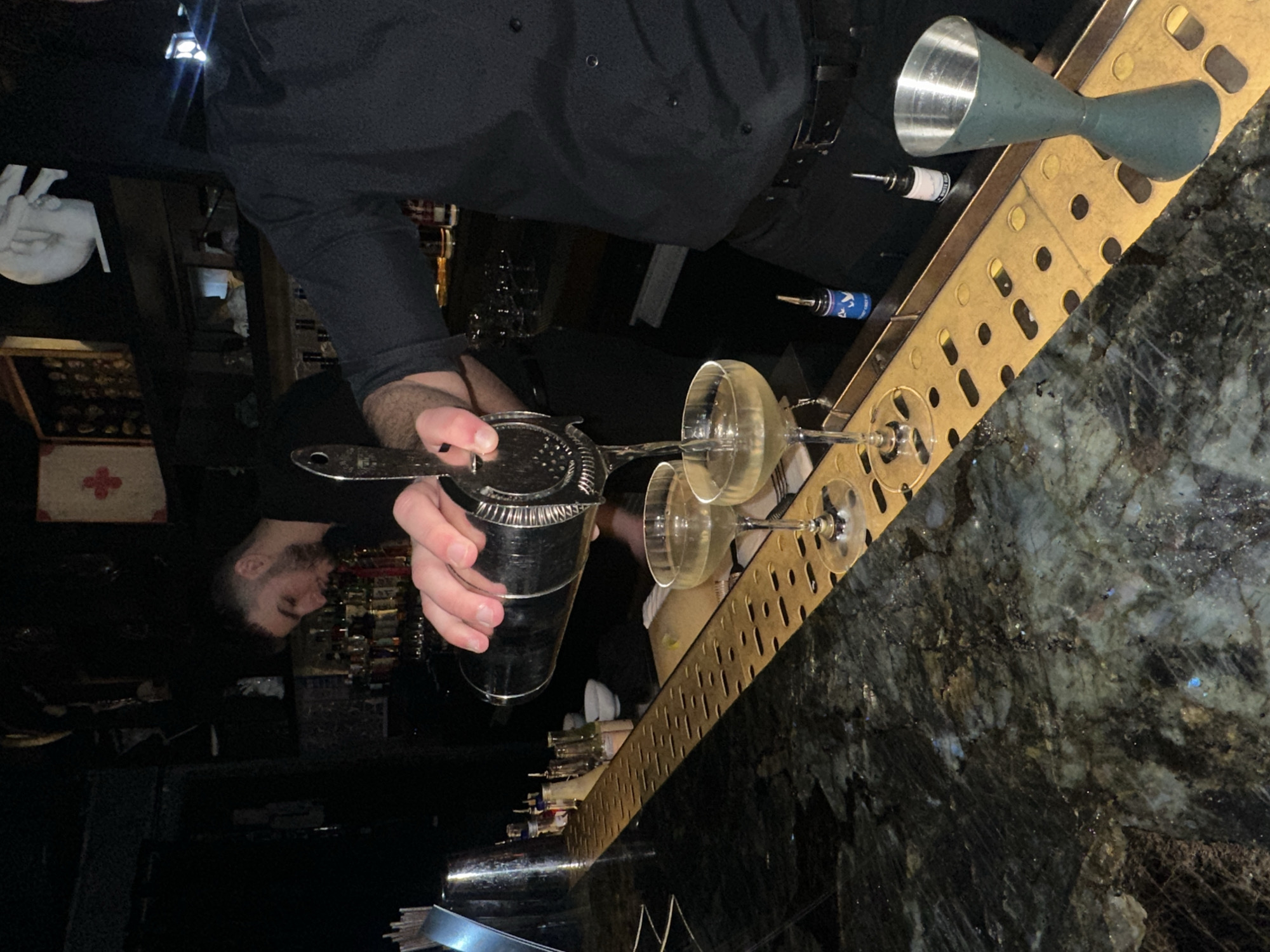 A bartender in London, United Kingdom, stands behind a bar, preparing cocktails. He is wearing a black shirt and apron. He holds a cocktail shaker in one hand, and a strainer in the other. He is pouring the contents of the shaker into a cocktail glass. The bar is made of a dark granite or marble, and has a metal bar top. There are bottles of alcohol and other bar equipment on the bar. The background is dark and out of focus, but it appears to be a bar or restaurant setting.