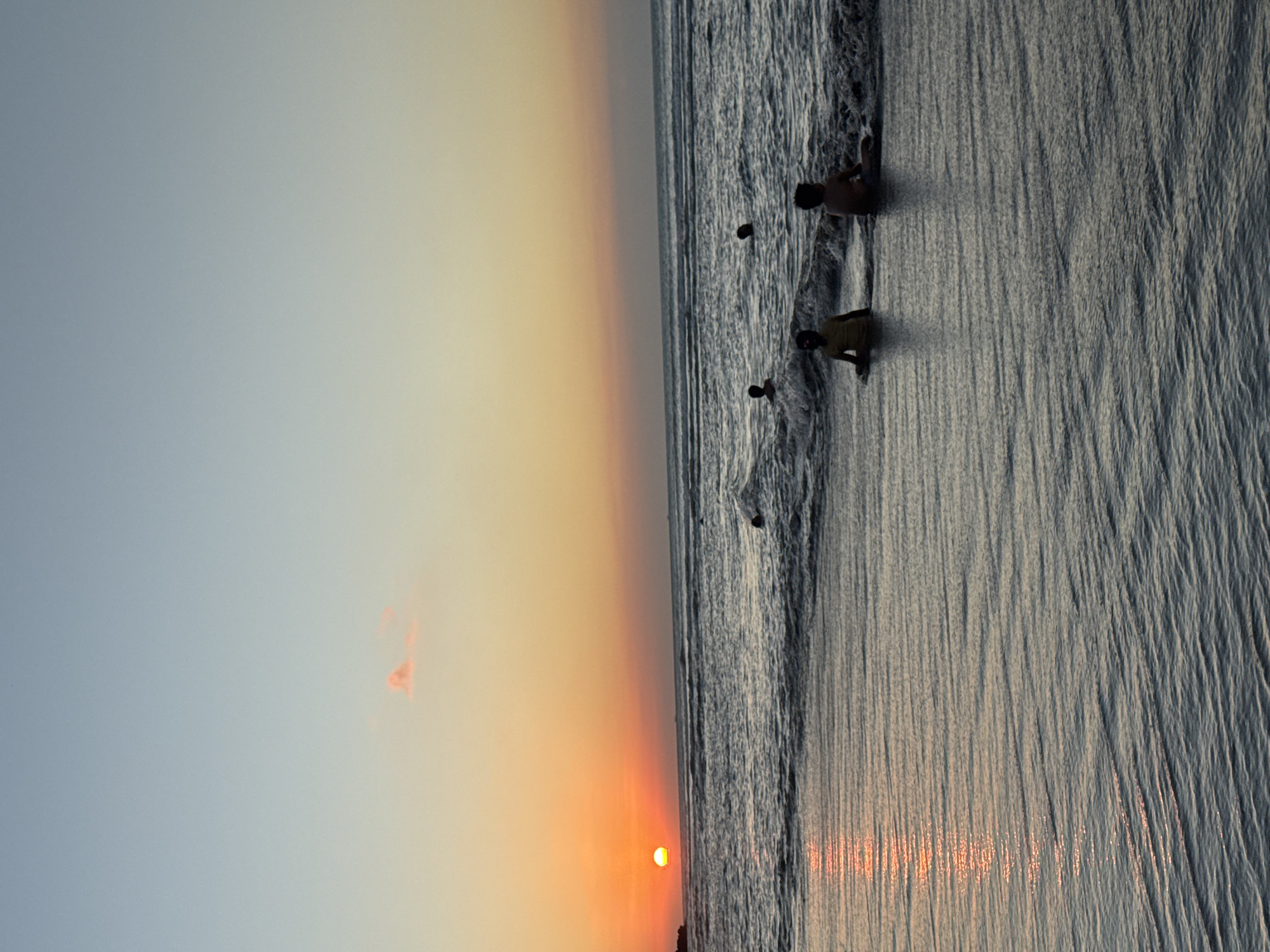 This media file depicts a serene sunset scene at a beach in Kuta Selatan, Indonesia. The sky, occupying the upper portion, transitions from a soft, pale blue-grey at the top to vibrant orange and warm yellow tones closer to the horizon. A small, faint reddish cloud is visible in the warmer part of the sky.
The sun, a bright orange-yellow orb, is setting low on the horizon, casting a brilliant glow. Its reflection creates a shimmering path of light across the ocean's surface and the wet sand in the foreground. The ocean, appearing in darker tones, shows gentle waves breaking near the shore. The wet sand is intricately textured with fine, parallel ripples left by receding waves, which also reflect the sunset's colors.
Several people are visible near the water's edge, engaging in leisurely activity. One individual stands in the shallow water, with their back to the viewer. Another person is seen kneeling or sitting closer to the shore, and a smaller figure, possibly a child, is also present near the water. A fourth, more distant figure can be faintly observed further out in the ocean. They appear to be enjoying the calm environment as the day ends.
The time of day is clearly sunset, highlighted by the low sun, the warm hues across the sky, and the reflective light on the water and sand. The overall mood of the scene is peaceful and tranquil. There is no visible text in the image.
