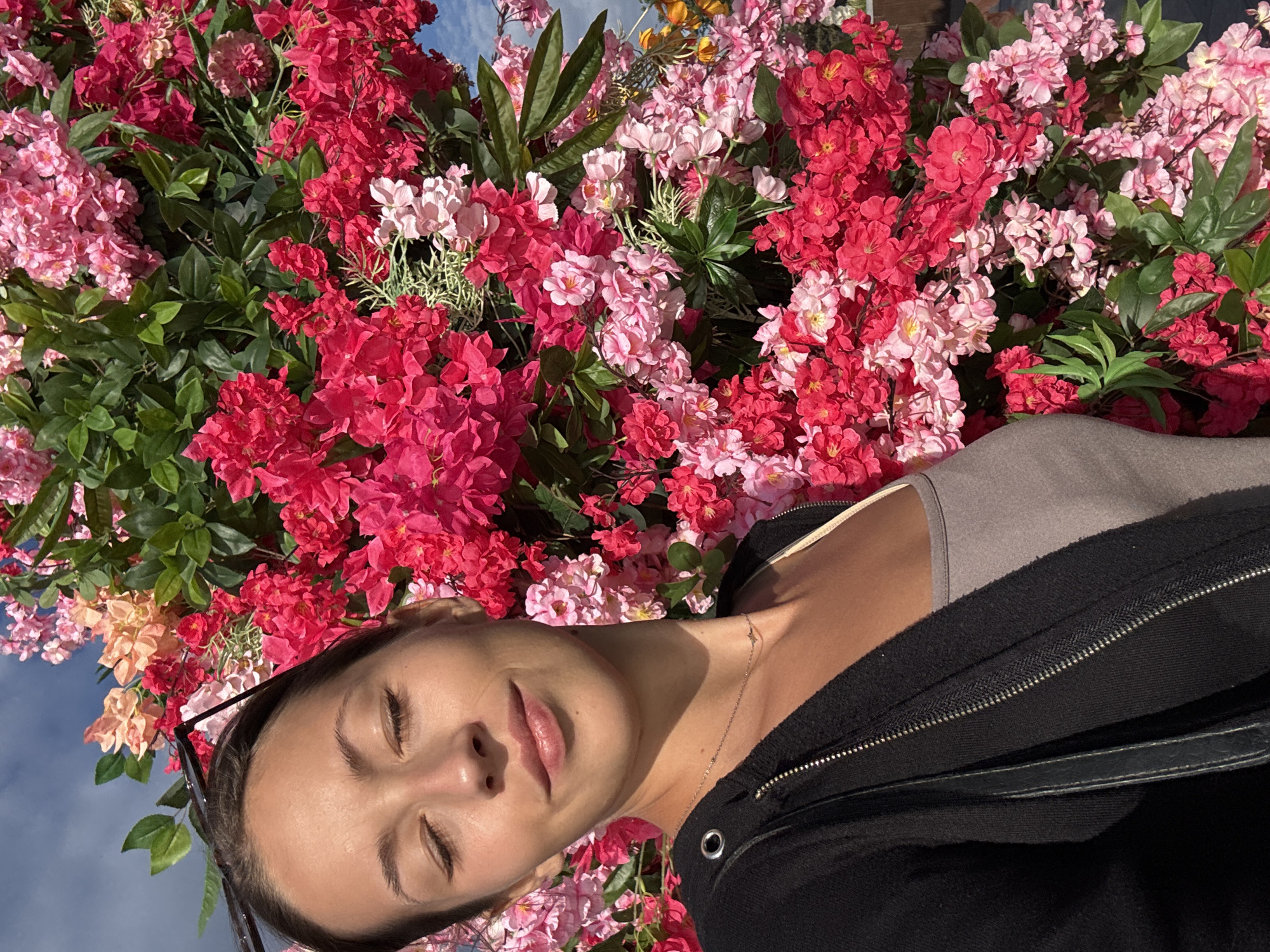 A young woman with closed eyes is lying beneath an array of pink and red flowers, with green leaves visible among the blossoms. She wears a gray top and a black jacket with a visible zipper. A necklace is visible around her neck. Sunglasses rest on her head near her hairline. The background shows a hint of blue sky. The location is Moscow, Russia.