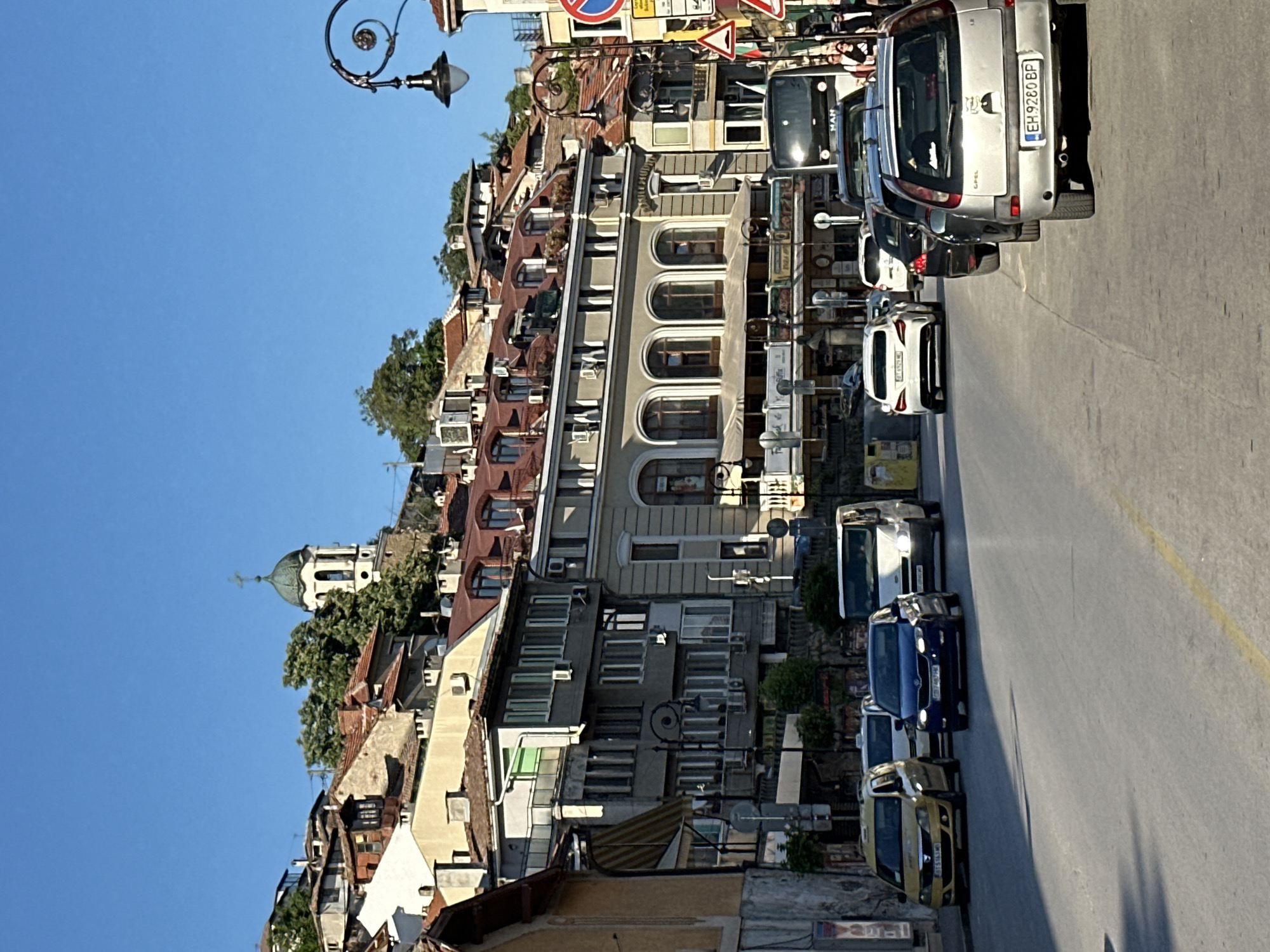 The image shows a street in Veliko Tarnovo, Bulgaria. The street ascends a hill, lined with buildings on the left and vehicles parked along the right side. A church dome is visible in the distance on the left. The buildings display a mix of architectural styles, including older structures with red-tiled roofs and more modern facades. The vehicles are parked close together, occupying much of the roadway. The sky is a clear blue.