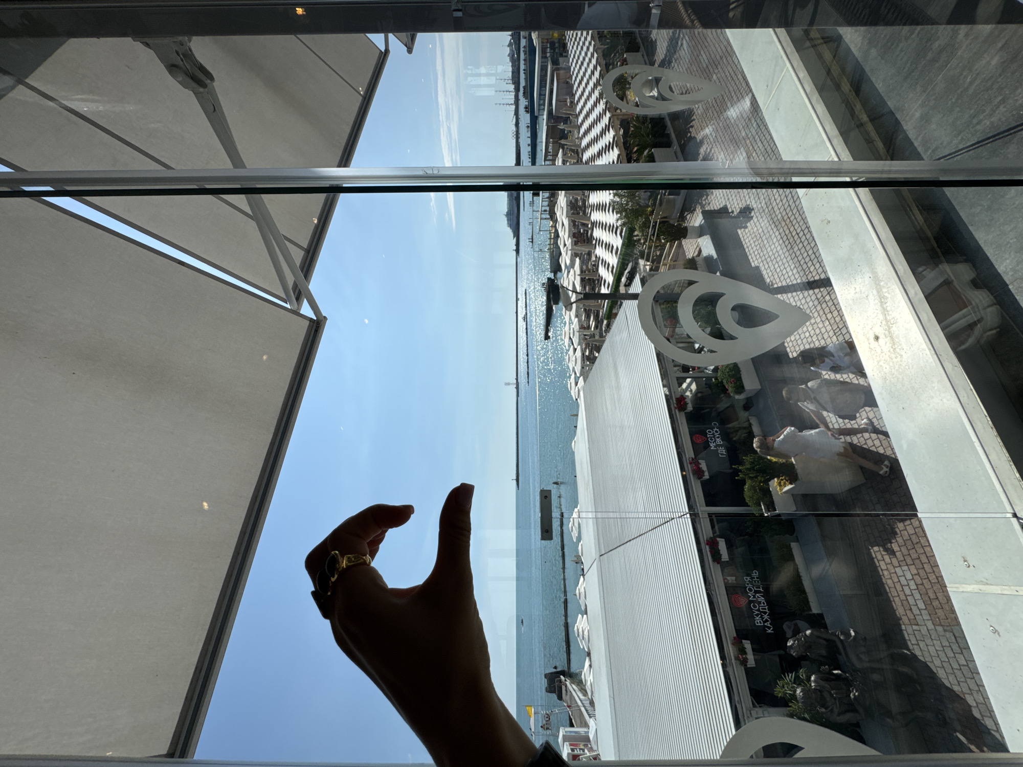 The image is taken from inside a building, looking out through a large window. A silhouetted hand with a ring on its index finger is forming a heart shape in the foreground, obscuring a portion of the view. Beyond the hand, a sunlit beach scene unfolds. Numerous white beach umbrellas are arranged on the sand, interspersed with patterned awnings. People are visible walking on the beach and near the water. In the distance, the sea stretches out, with boats and a large ship on the horizon under a clear blue sky. The location appears to be a coastal establishment in Sochi, Russia, possibly a restaurant or cafe, given the visible signage in Russian that reads "ВКУС МОРЯ КАЖДЫЙ ДЕНЬ" (Taste of the Sea Every Day) and "МЕСТО ГДЕ ВКУСНО" (A Place Where It's Tasty). The reflections on the glass suggest an indoor setting with artificial lighting.