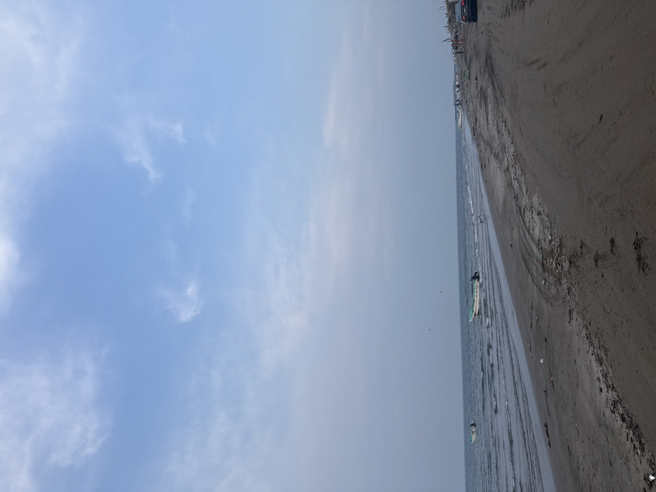 The image displays a wide, ground-level view of a beach in Antón Lizardo, Mexico, under a light blue sky with scattered, wispy white clouds. The ocean, characterized by blue-grey water and small waves, occupies the left side of the frame. Two small, white boats with light green trim are positioned in the shallow water near the shore in the mid-ground, one featuring a visible outboard motor. The beach itself is composed of dark grey sand and dirt, extending from the foreground into the distance on the right, and exhibits multiple vehicular tire tracks. Scattered debris is present along the waterline and on the sand. In the far distance on the right, a dark-colored sedan is parked on the sandy path, and several indistinct human figures are visible on the beach. Beyond these figures, additional small boats are either in the water or pulled onto the sand, adjacent to what appear to be rudimentary structures constructed from wood and tarpaulins. A single piece of metallic debris is partially submerged near the water's edge in the mid-ground.