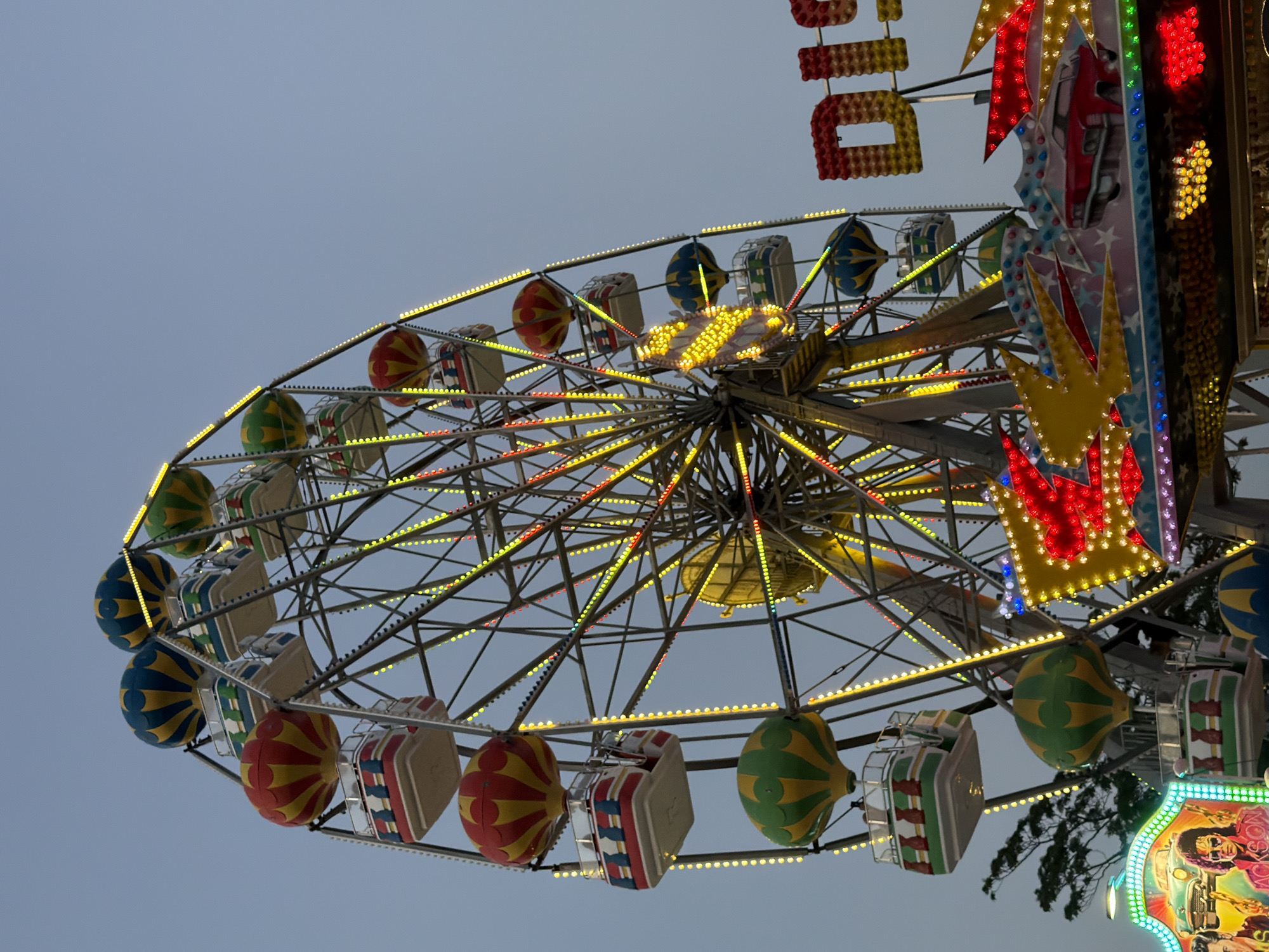This media file captures a low-angle view of a Ferris wheel at what appears to be an amusement park or fairground. The sky is a pale, overcast gray, suggesting it could be twilight or a heavily clouded day. The Ferris wheel is adorned with numerous colorful lights, primarily yellow and red, which are beginning to glow against the dim sky. Various gondolas are attached to the wheel, some appearing as pods with red and yellow striped patterns, while others are more box-like with white and red checkered designs. Some gondolas are also decorated with green and yellow patterns.
To the right of the Ferris wheel, a section of another amusement ride is visible, brightly lit with red, yellow, and green lights. This ride features decorative elements, including what look like stylized yellow stars and red diamond shapes. There is also a portion of a sign with red and yellow block letters that appears to spell out "DICK". Further to the right, a vibrant, colorful sign with cartoonish depictions of people and text is partially visible at the bottom right corner.
The overall impression is one of a festive atmosphere at an amusement park, possibly as evening approaches or during overcast conditions. The presence of numerous lights on the rides suggests it is operational. No people are clearly visible within the gondolas or on the ground. The location context provided is Varna, Bulgaria, which might imply this is a local fair or amusement park in that city.