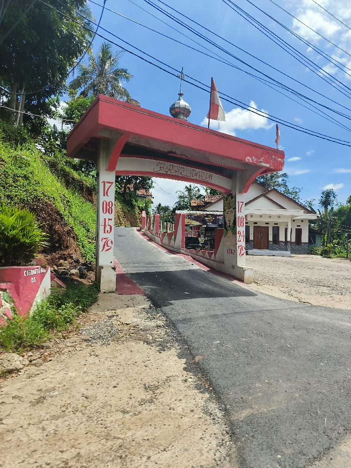 A red and white archway with the words "Selamat Datang" (Welcome) on it stands over a paved road. There is a small white building on the right side of the archway. The road leads uphill to a small village in Cipatat, Indonesia. The coordinates are -6.778 latitude, 107.437 longitude. There are trees lining the road on the left side of the archway. There is a red and white sign on the left side of the archway that says "Cibenten 01". The archway is decorated with numbers and designs. There are a few flags hanging from the archway. There is a small dome on top of the archway. There are a few people walking in the distance. The sky is blue with white clouds. The scene is well lit.