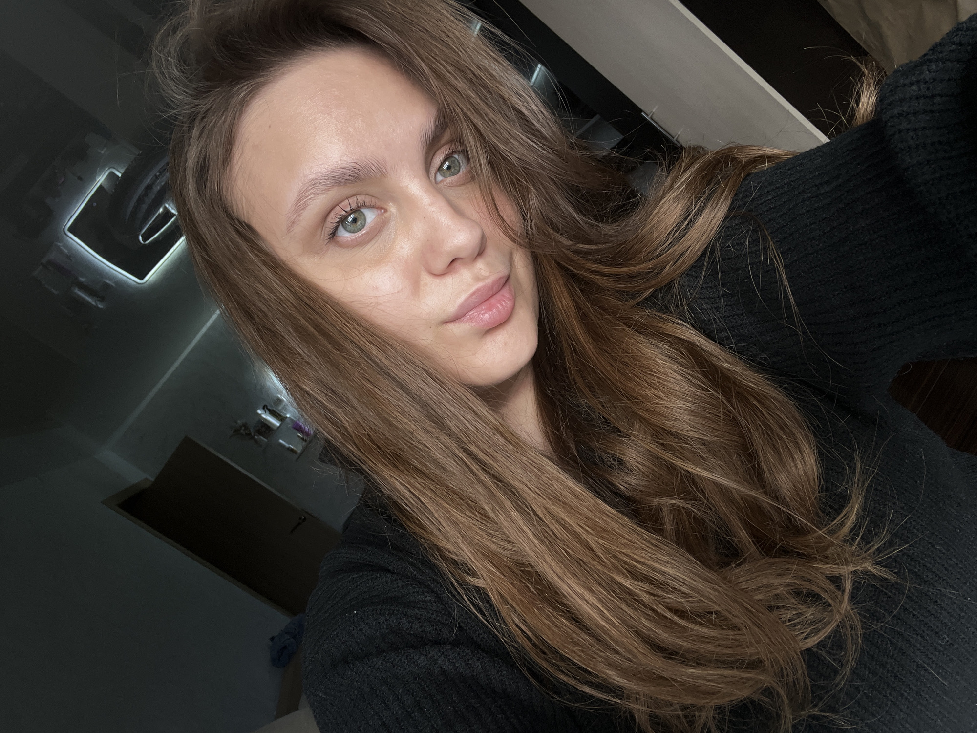 A fair-skinned woman with long, brown hair is centered in the frame, taking a selfie. She is wearing a black sweater. Her eyes are light green. The background is blurry, but reveals interior details of a room, including a light fixture, a door, and a wall. The image appears to be taken indoors. It is impossible to determine the exact location in Chaikovsky, Russia without further context or information.