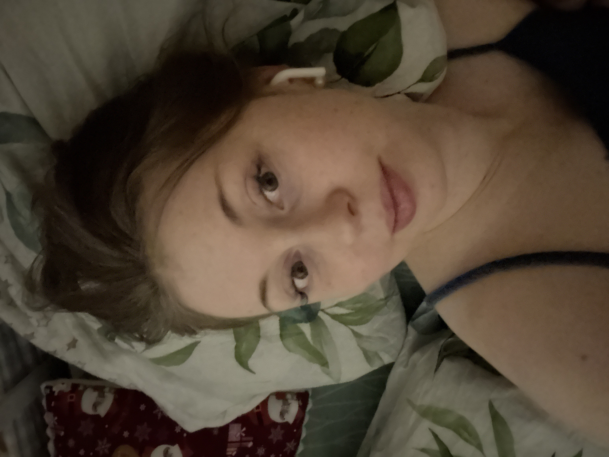 The image shows a close-up, slightly angled shot of a young person, likely a woman, lying down. Her head is resting on a pillow with a light-colored background and a pattern of green leaves. She has light skin, brown eyes, and dark brown or auburn hair that appears somewhat disheveled.
A white wireless earbud is visible in her right ear. Her expression is neutral, with her gaze directed towards the camera. Her lips are slightly parted, revealing a soft pink color. She is wearing a dark-colored strap of clothing, possibly from a tank top or nightwear, visible on her right shoulder.
To her left, another pillow or blanket can be seen, featuring a red background with white or light-colored patterns, which could be festive or holiday-themed. The lighting in the room appears dim, suggesting it might be evening, early morning, or simply a softly lit indoor environment. The overall setting appears to be a bedroom, with the person relaxing in bed. There is no visible text in the image.