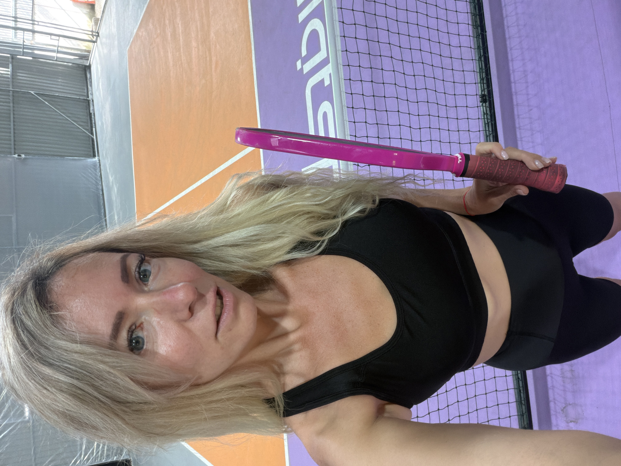 The image shows a woman holding a pink paddle, standing on a purple court with an orange area behind her. The court appears to be an indoor sports facility, possibly for padel or a similar racket sport, with a net in the foreground. The woman has blonde hair and is wearing a black sports bra and black shorts. The background suggests an industrial or warehouse-like setting with corrugated metal walls. The lighting is artificial, likely from overhead sports lights. No specific landmarks or details indicate the location in Da Lat, Vietnam.