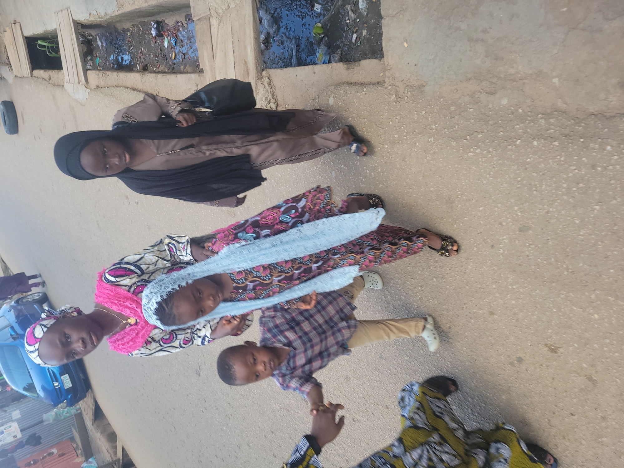 In Jos, Nigeria, a group of four children walks along a paved road, with a drainage ditch on the right side of the image. The children are dressed in traditional attire. The child in the front is wearing a light blue headscarf and a patterned dress. To her left, a young boy in a plaid shirt and tan pants is being held by a woman whose arm and patterned dress are visible on the far left. To the right of the front child is an older girl in a black hijab and a brown dress, carrying a black purse. Behind her, another girl is partially visible, wearing a pink and white patterned outfit and a headscarf. The road is a light gray asphalt, with some cracks and pebbles. The drainage ditch is filled with murky water and debris, with wooden planks covering parts of it. In the background, a blue car is parked on the left side of the road, and a few buildings are faintly visible. The overall scene depicts a typical street view in Jos, with elements of daily life and urban infrastructure.