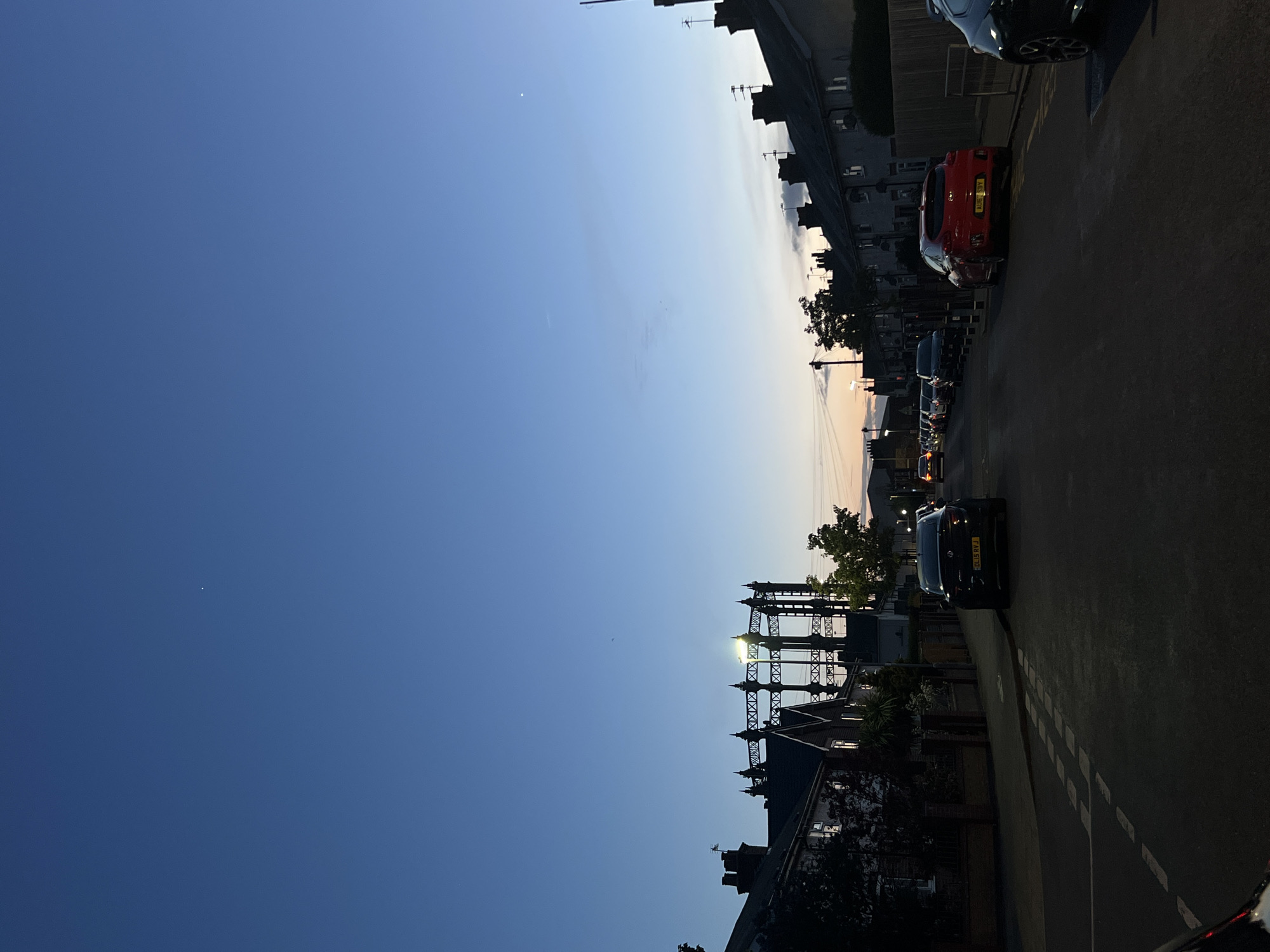 The image depicts a street scene in Great Yarmouth, United Kingdom, taken at dusk or dawn. The sky transitions from a deep blue to a lighter gradient near the horizon. Silhouetted rooftops and chimneys line the lower right edge, partially obscuring a large, ornate gas holder structure. A row of parked cars occupies the right side of the narrow street, including a red hatchback with license plate visible as "AI18 SBY", and a black car with a license plate partially visible as "FA18 NVJ". Streetlights cast a soft glow, illuminating the road surface with white line markings. The overall ambiance is quiet and residential.