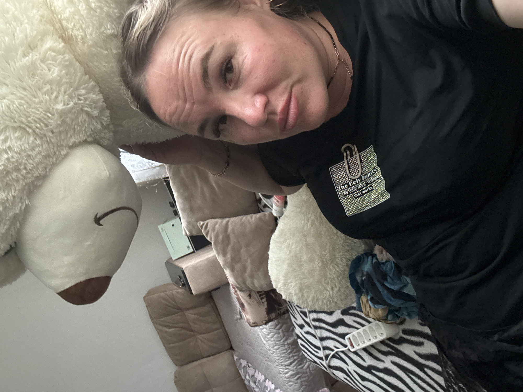 A woman lies on a couch in a home in Pozdyshki, Russia, with a large, fluffy white teddy bear behind her. She is wearing a black t-shirt with a bejeweled patch that reads "The Holy Grail" and other text. She has blonde hair pulled back and is wearing hoop earrings and a necklace. The couch has brown and tan cushions, and there is a zebra-print throw pillow visible. A power strip with multiple outlets is also on the couch. The overall impression is of a casual, domestic setting.