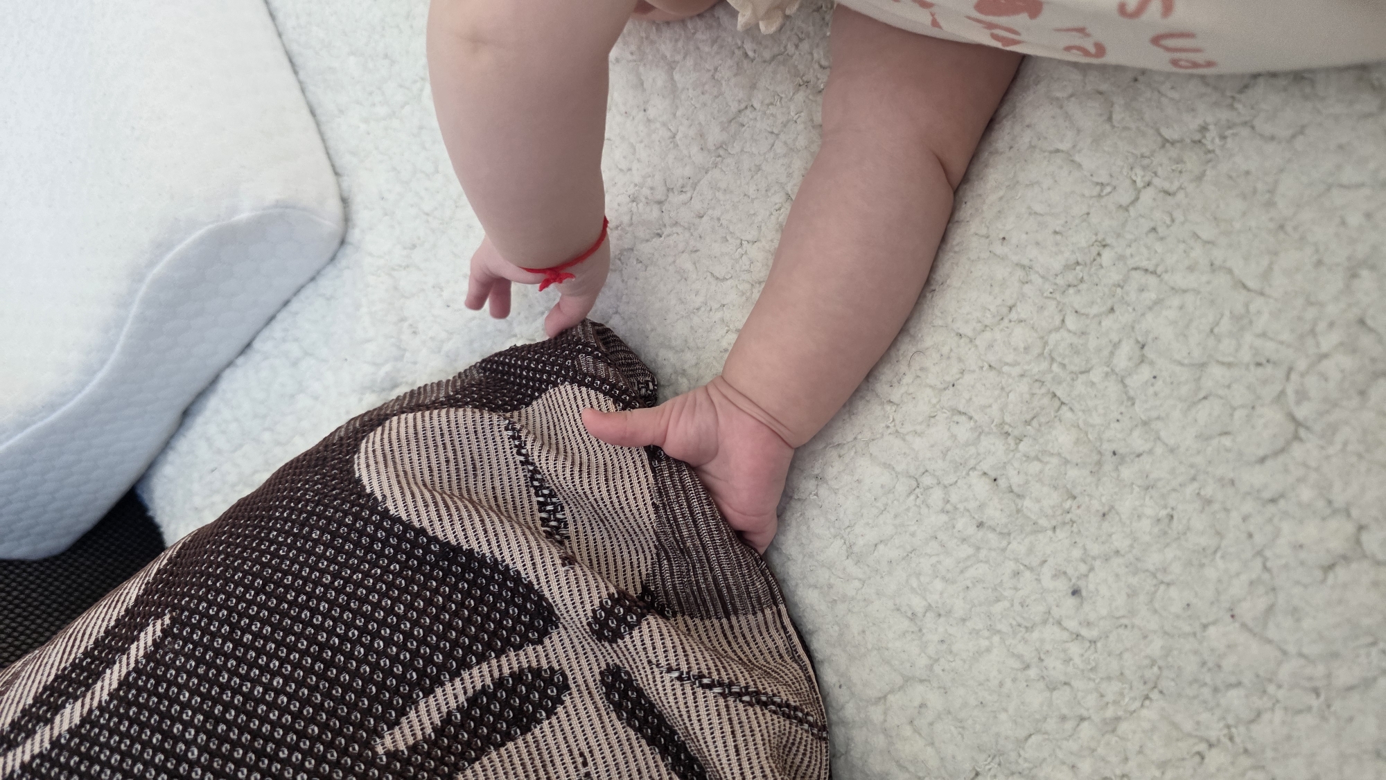 A baby's right arm and hand are visible on the right, with the hand grasping a brown and beige patterned pillow. The baby wears a light-colored garment with a print on it. The left arm is also visible with a red string bracelet. A white pillow is on the left. The background consists of a textured, light-colored surface, possibly a blanket or rug. The image does not provide any information to confirm the location in the city of Blagoevgrad, Bulgaria.