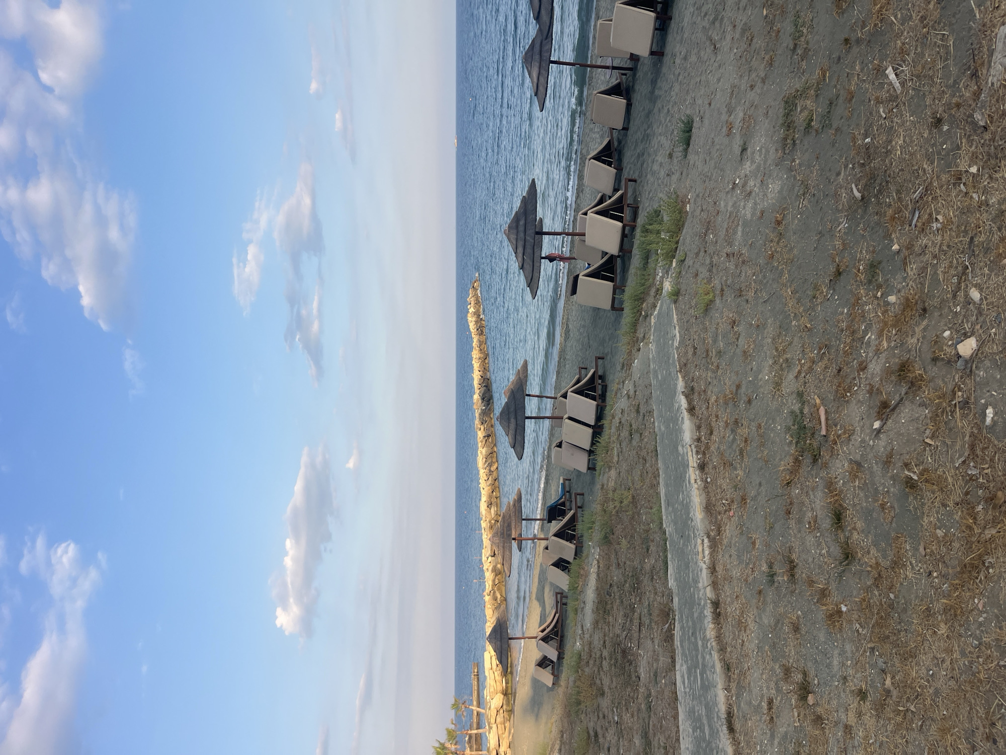 The image depicts a serene beach scene in Agios Tychonas Community, Cyprus. The foreground is dominated by dry, sandy ground with sparse vegetation and a grey paved pathway. In the middle ground, several rows of empty lounge chairs and thatched umbrellas are arranged on the dark sand, facing the calm blue sea. A stone breakwater stretches across the water in the background, with a single person visible near the breakwater. The sky is a clear blue with scattered white clouds, and a distant boat is visible on the horizon. The overall impression is one of tranquility and a resort setting.