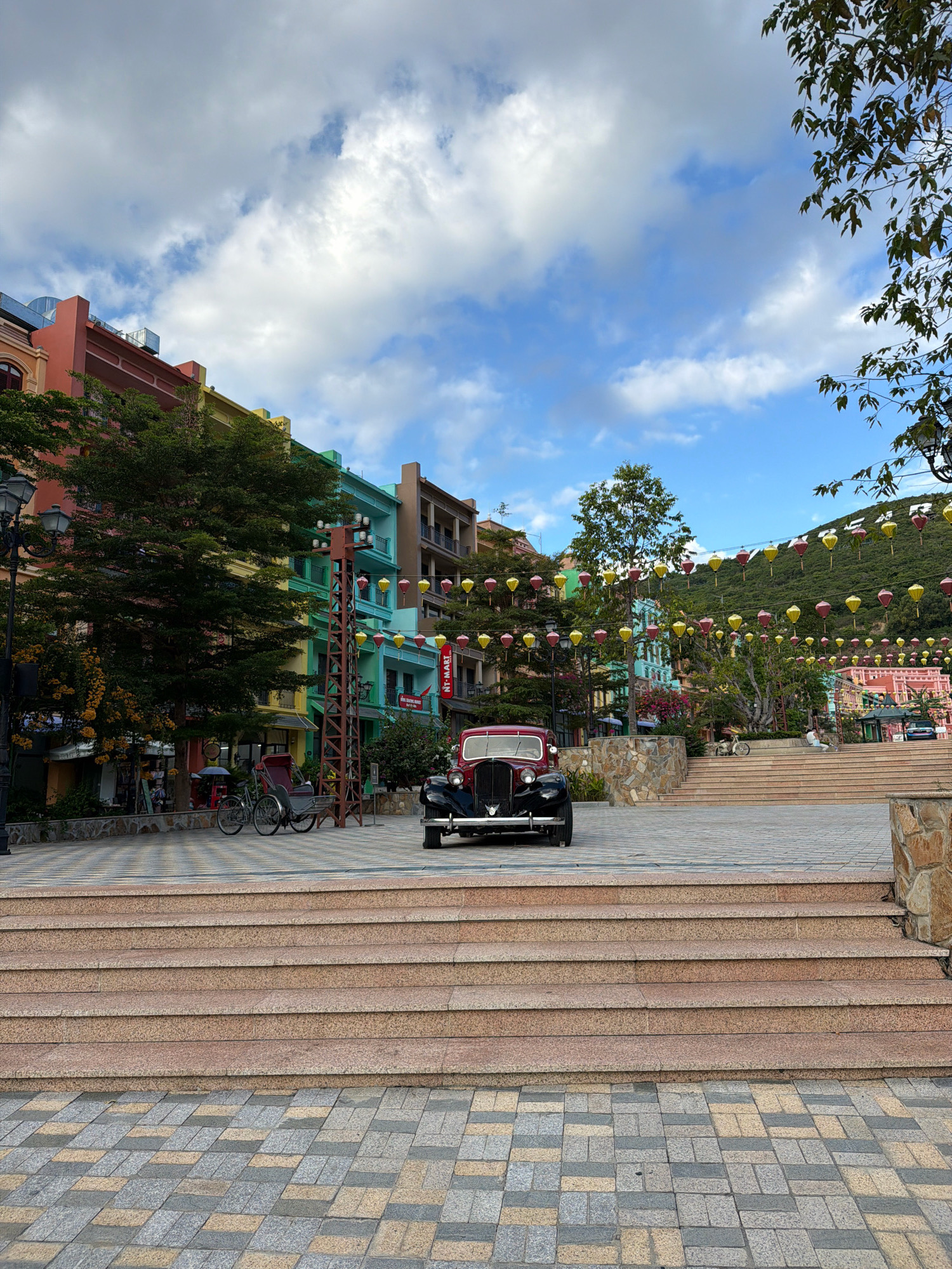 A vintage red car is parked in a paved plaza in front of a set of stone stairs in Nha Trang, Vietnam. The plaza is lined with colorful buildings and decorated with strings of lanterns. Lush green trees and foliage are visible throughout the scene. The sky above is a mix of blue sky and white clouds. The pavement in the foreground is made of interlocking stone tiles in shades of gray, yellow, and beige. The stairs are made of pinkish-brown stone. A bicycle rickshaw is parked to the left of the car, and a lamp post is visible behind it. The buildings are painted in various bright colors, including pink, orange, yellow, green, and blue. The background shows a hillside covered in trees and more lanterns.