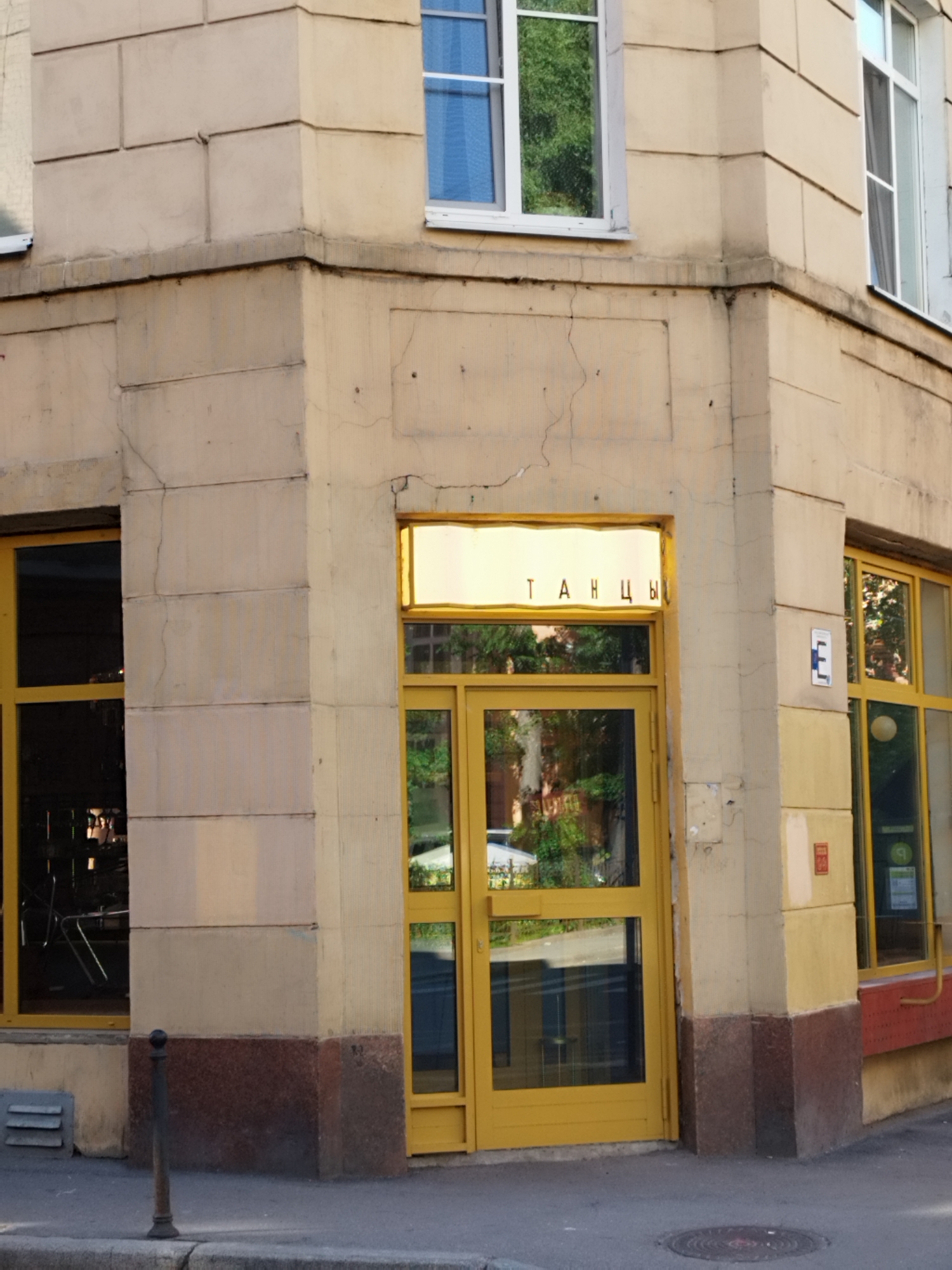This is a photograph of the corner of a light beige, stucco-clad building, likely in Saint Petersburg, Russia, given the Cyrillic text. The building features several large windows with white frames, some reflecting green trees. A prominent yellow door with glass panels is visible on the corner, leading into the building. Above the door, a rectangular sign with yellow lighting displays Cyrillic text in black letters: "ТАНЦЫ," which translates to "DANCES." To the right of the door, another yellow-framed window shows a reflection of trees and greenery. A small blue and white sign with the letter "E" is mounted on the wall near this window. The base of the building is adorned with a dark red, granite-like material. A thin, dark metal pole stands on the sidewalk in front of the left side of the building, which also has a louvered vent near its base. The scene appears to be captured during daylight, possibly late afternoon, given the warm lighting and shadows. The overall impression is of an urban storefront.