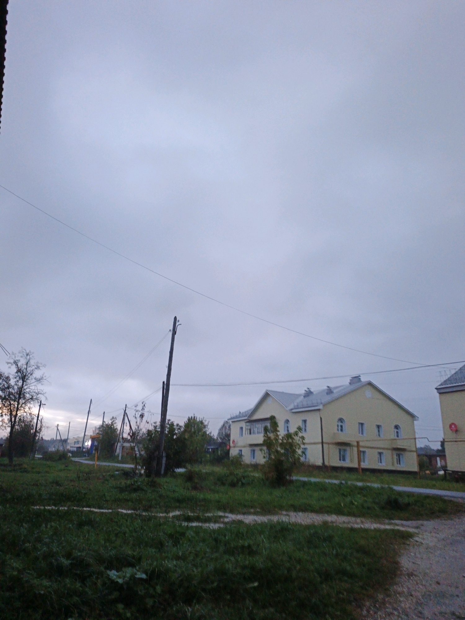 The image captures an overcast sky over a village scene in Medvedyovka, Russia. In the foreground, a grassy field stretches across the bottom half of the frame, with some areas of uneven terrain and a faint path. Several wooden utility poles with visible wires are scattered throughout the middle ground, including a prominent one in the center-right. Behind the field and poles, a light yellow, two-story building with a grey roof and numerous windows is visible. This building appears to be a residential structure, possibly an apartment building, with arched windows on the upper floor. To the right of this building, another similar structure, partially obscured, is also present. Trees and foliage are scattered across the landscape, some appearing bare and autumnal. The overall atmosphere is subdued, consistent with a cloudy or twilight day. No people are discernible in the image.