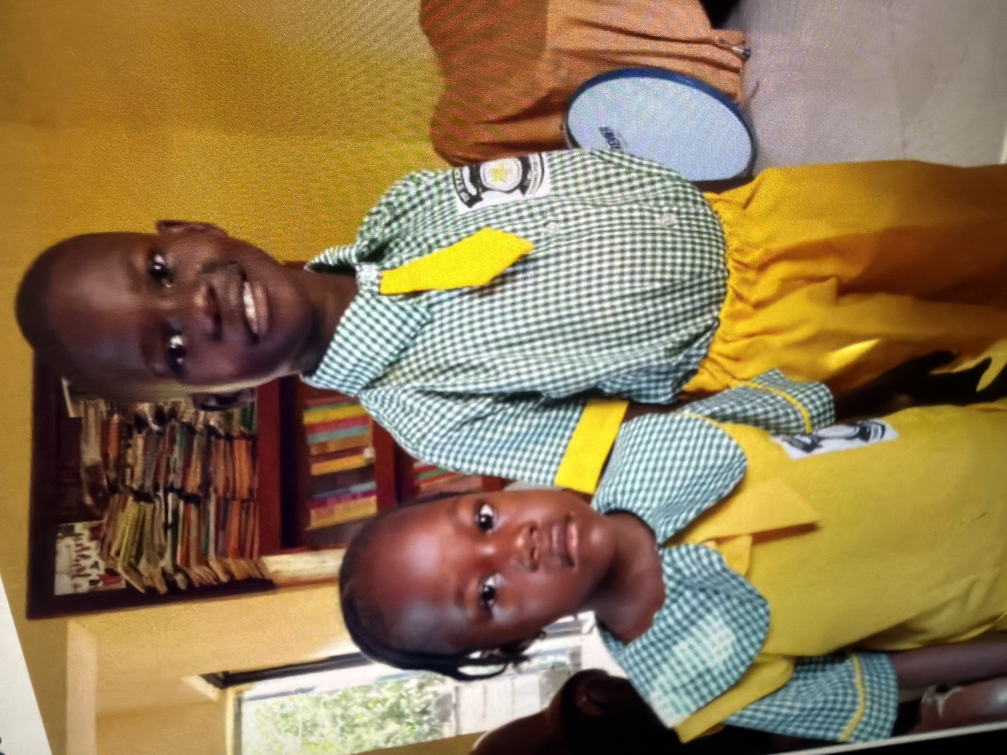 Two children, a boy and a girl, stand in front of a bookshelf filled with books. The boy, on the right, smiles at the camera and wears a green and white checkered shirt with a yellow tie and yellow pants. He has a patch on his shirt sleeve. The girl, on the left, looks seriously at the camera and wears a yellow dress with a green and white checkered collar and sleeves, also with a patch on the sleeve. The room has yellow walls and a window in the background. To the right of the children, a circular object with a blue rim is visible, and behind it, an orange draped fabric. The overall impression is a casual indoor setting, possibly a home or a school.