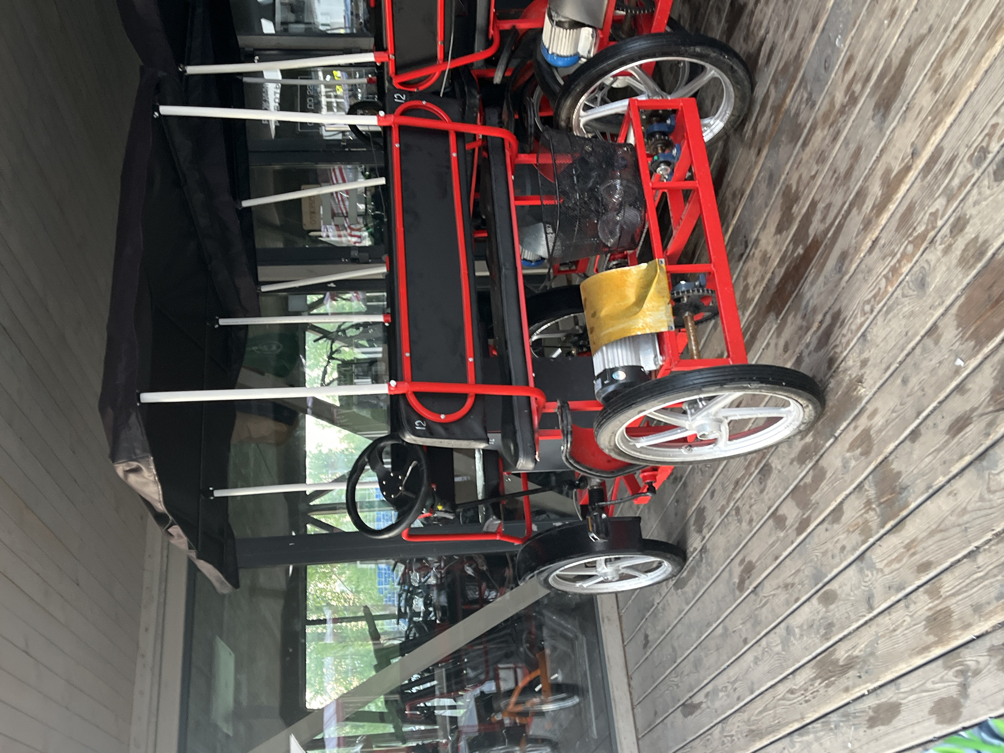 A tandem quadracycle is parked on a wooden deck adjacent to a building with large glass windows. The quadracycle is red and black with white wheels. A dark canopy is over the quadracycle. The reflection in the window shows several bicycles parked inside the building. The location is Krasnoyarsk, Russia.