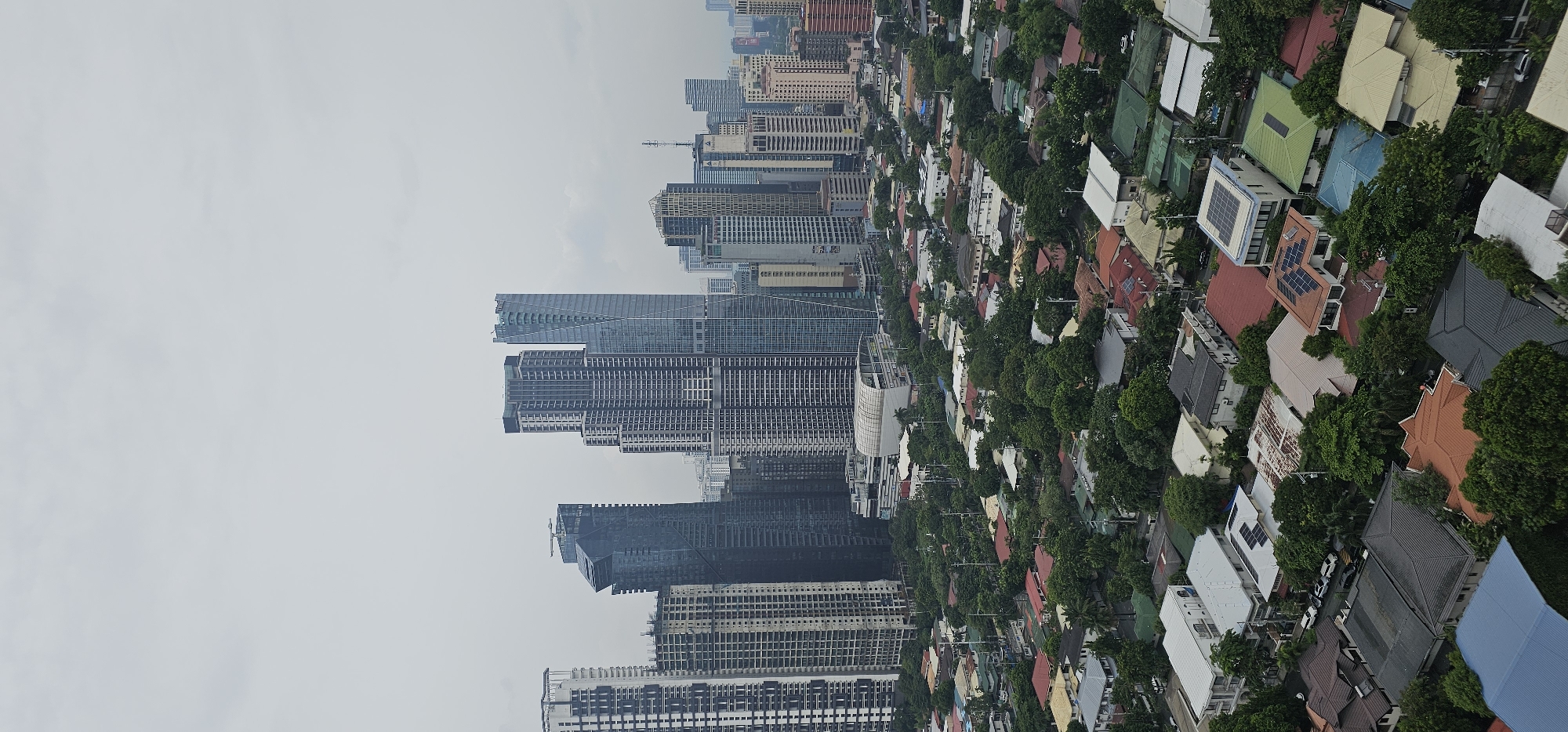 This is an aerial view of a city skyline in Makati, Philippines, featuring a dense urban landscape. In the background, a cluster of modern skyscrapers with varying architectural designs dominates the scene under an overcast sky. The buildings are characterized by their height, glass facades, and intricate structures.
In the foreground and midground, a residential area with a multitude of houses is visible. These houses are densely packed, showcasing a variety of roof styles and colors, including red, gray, and green. Many of the houses have trees surrounding them, creating a green canopy that partially obscures the roofs. Some houses are equipped with solar panels. The streets are narrow and appear to be lined with parked vehicles. The overall impression is one of a developed urban area with a mix of high-rise commercial buildings and lower-density residential neighborhoods.
