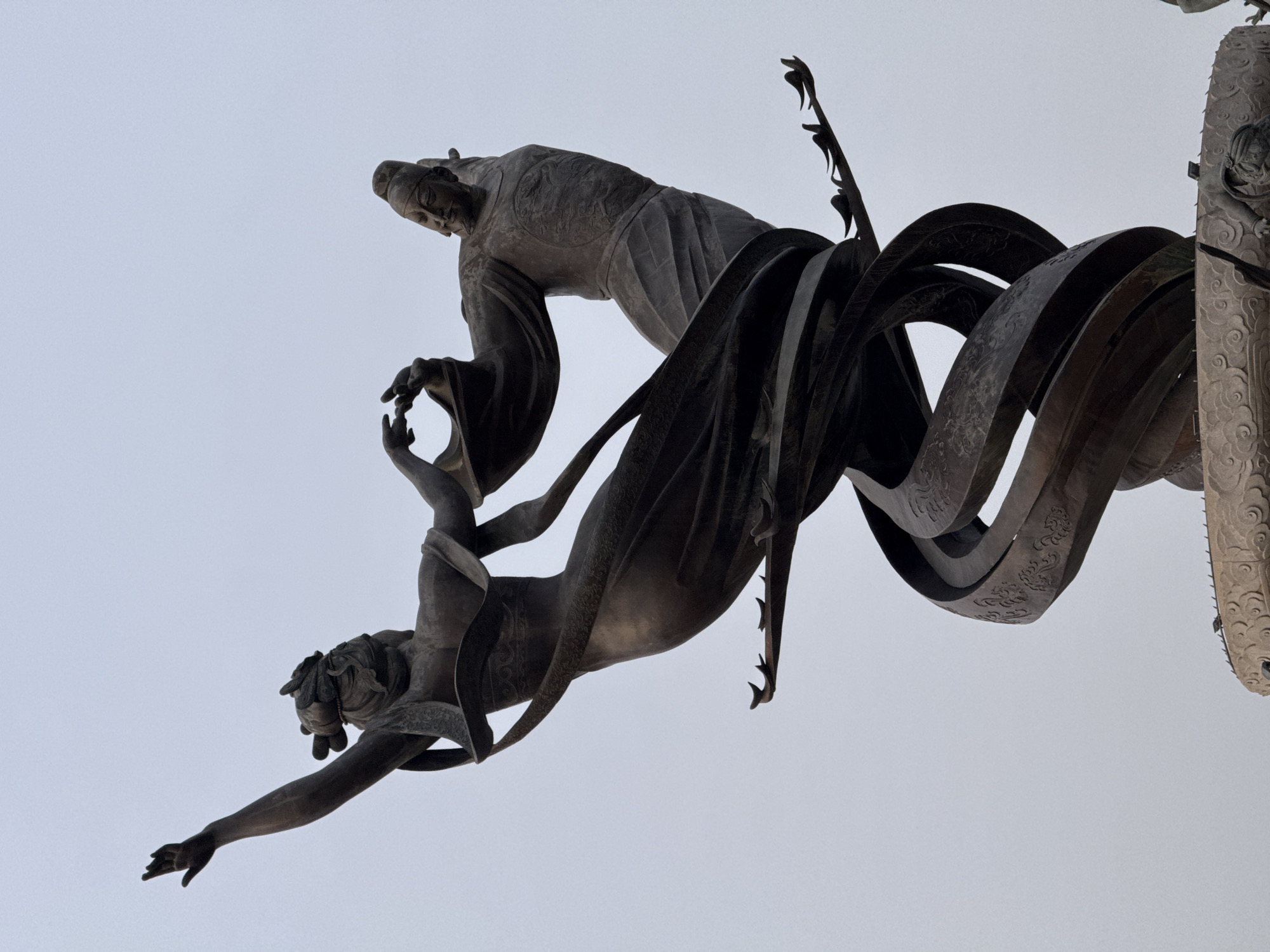 The image depicts a bronze sculpture, specifically a component of the "Long Mark" sculpture located at the Lintong Museum in the Lintong District, China. The sculpture features a standing figure in traditional Chinese attire grasping the hand of another figure below, who is reaching downwards. The figures are bronze and stand against a light sky. The sculpture also incorporates swirling, metallic ribbons with decorative patterns. To the right, a partially visible cylindrical column with carved embellishments is present.