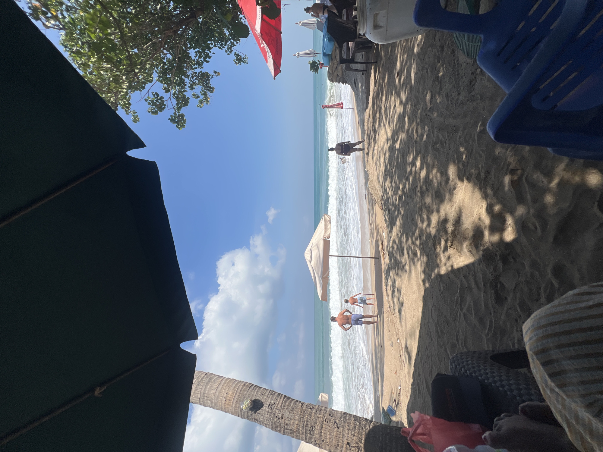 The image depicts a tropical beach scene in Kuta, Indonesia, on a sunny day. In the foreground, partially visible, are the legs of a person resting on a striped fabric, with a woven chair and a red bag nearby. The sand is covered in dappled shade from unseen foliage.
Further back, two people, a man and a child, stand on the wet sand near the water's edge, facing the ocean. The man is wearing blue swim trunks, and the child is also in swim attire. The waves are breaking gently on the shore.
Several beach umbrellas are visible across the scene. A large, dark green umbrella is prominent in the upper portion of the image, with a portion of a palm tree to its left. To the right, a red umbrella stands over a table with chairs and sandbags. In the background, more white umbrellas dot the beach, and the turquoise ocean stretches to the horizon under a bright blue sky with scattered white clouds. The overall atmosphere is relaxed and indicative of a day at the beach.