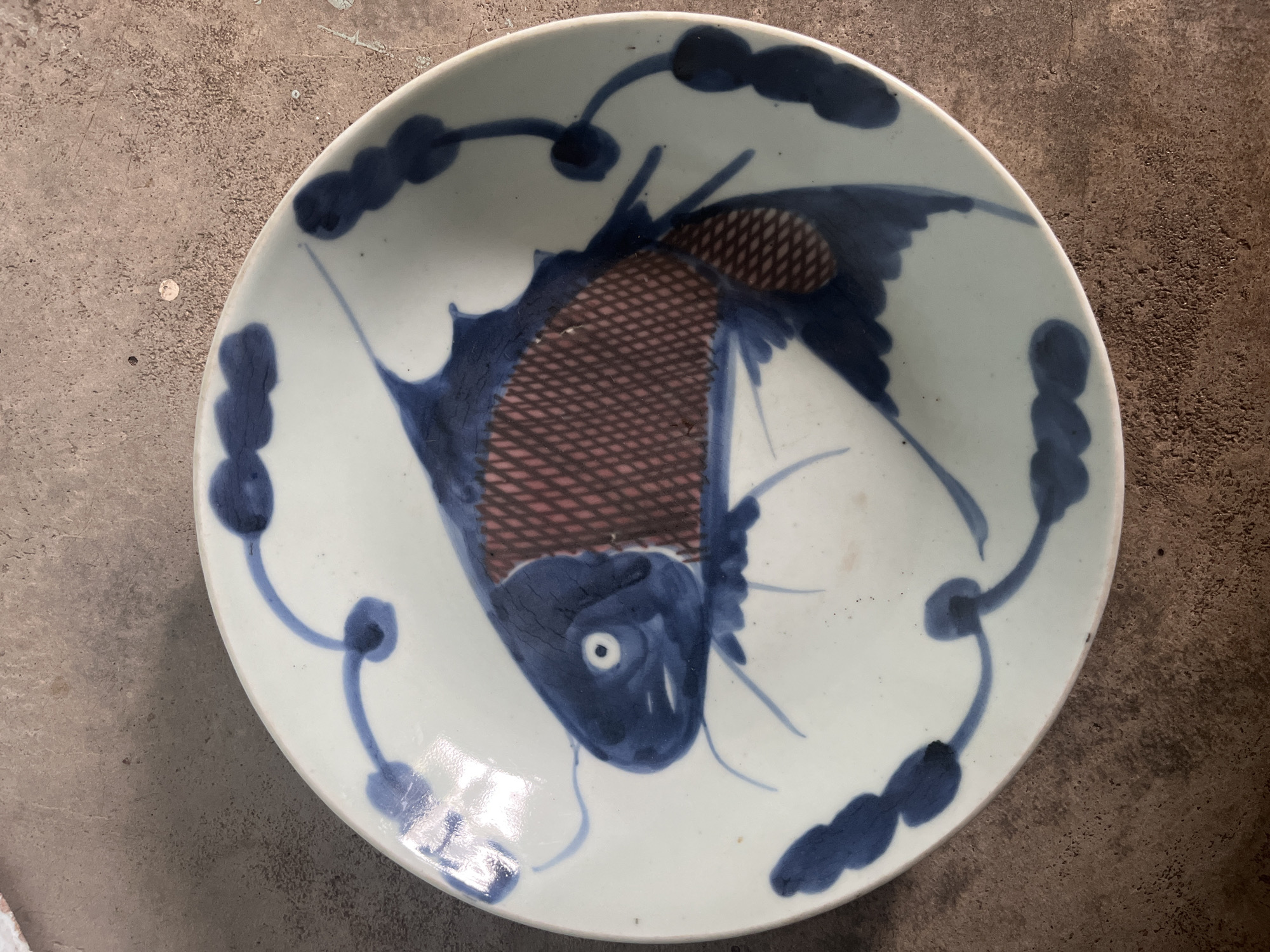 A close-up, overhead shot displays a white ceramic plate with a blue and reddish-brown painted fish design. The fish is depicted in a stylized manner, with blue for its head, fins, and tail, and a cross-hatched reddish-brown pattern for its scales. The plate is adorned with blue cloud-like or floral motifs around its rim, connected by delicate blue lines. The plate rests on a textured, concrete-like surface, with variations in color and tone suggestive of a weathered or outdoor environment. The lighting casts subtle shadows and highlights on the plate's glossy surface, emphasizing its curvature and the painted details. There is no indication of the city of Meliau Hulu, Indonesia, in the image.