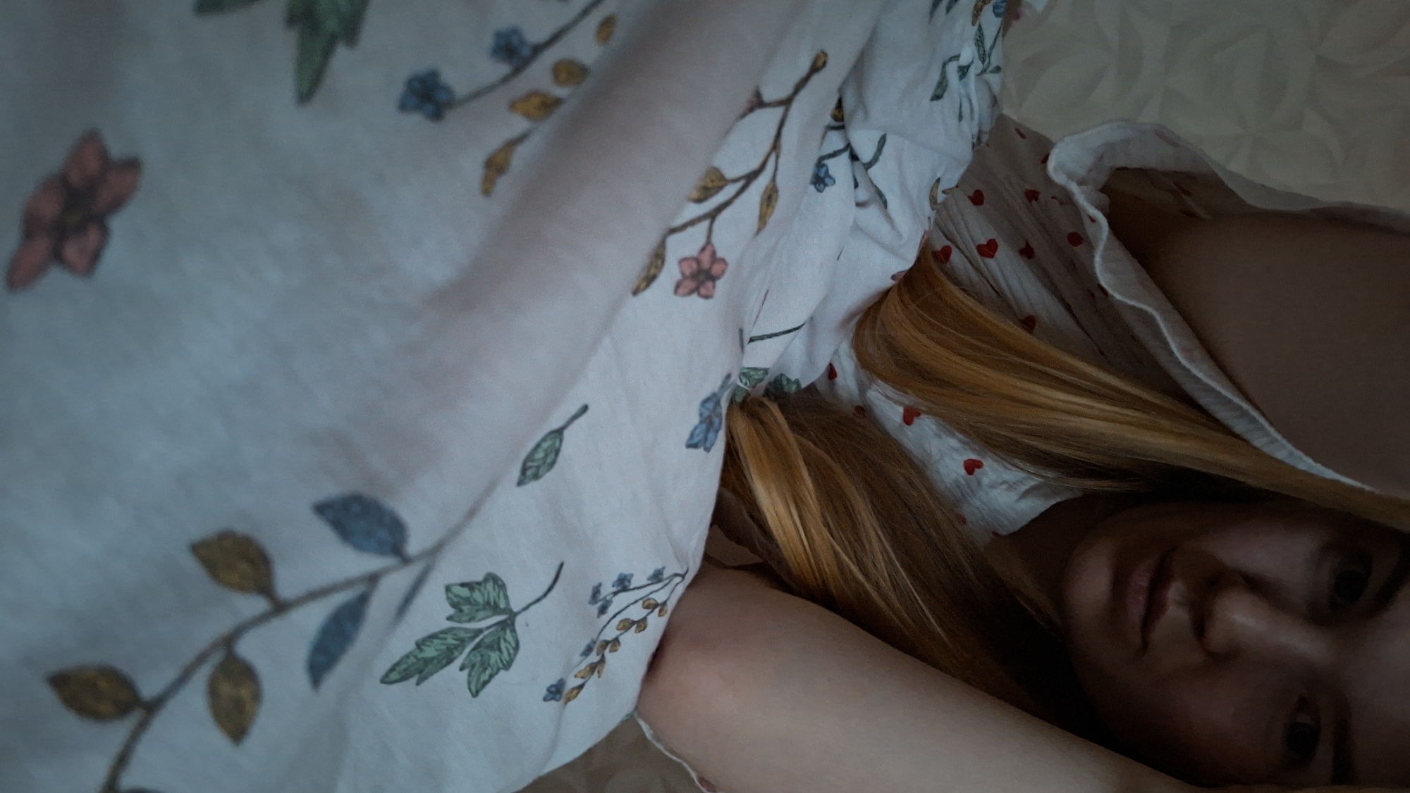 A woman with blonde hair is lying in bed, partially covered by a floral-patterned sheet. She is wearing a white garment with red hearts. Her face is visible, and she appears to be looking at the camera. The environment is indoors, likely a bedroom. There is no location given in the image.